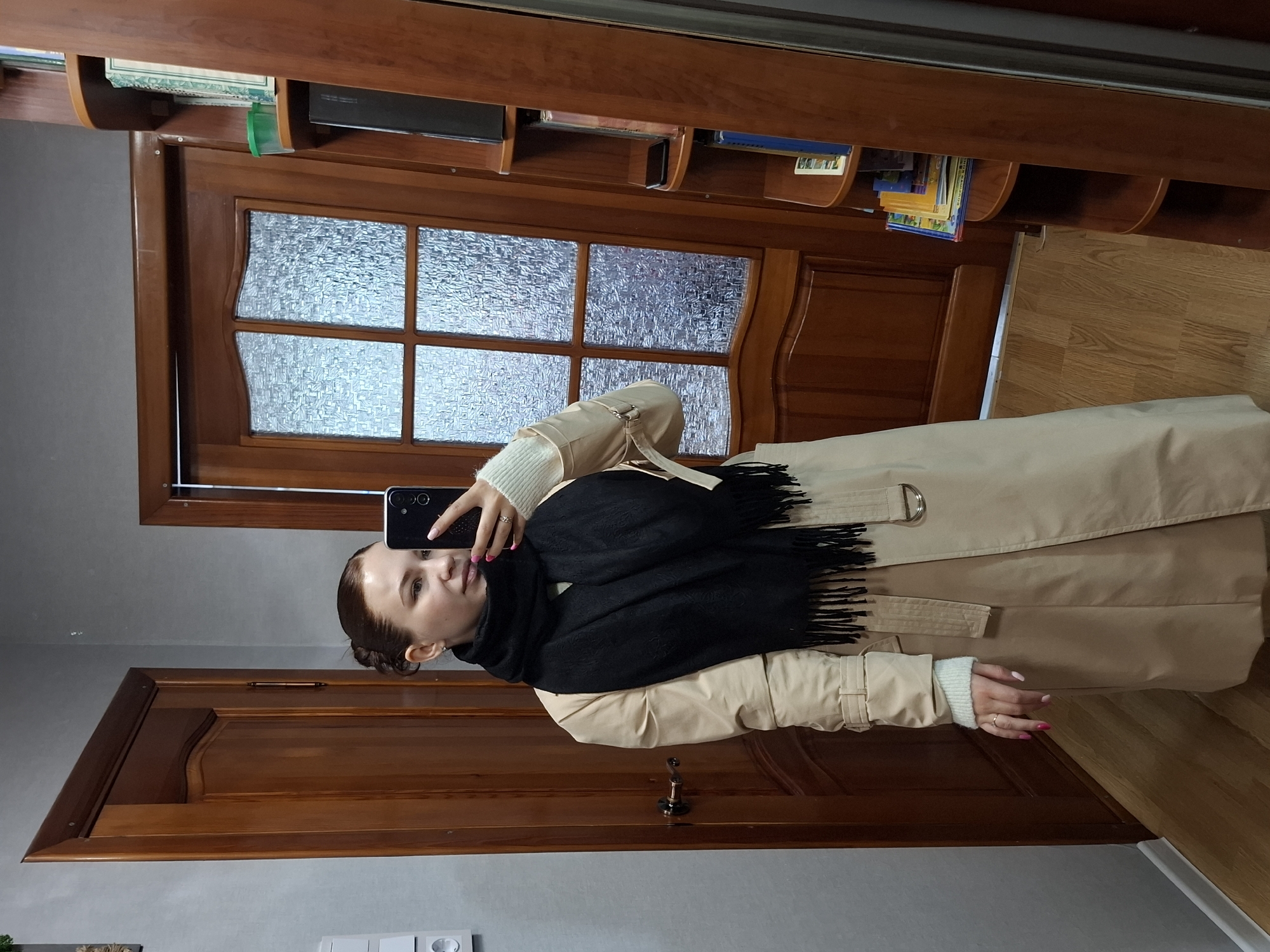 A woman is taking a selfie in front of a mirrored door in what appears to be a residential setting in Chaikovsky, Russia. She is wearing a light tan trench coat over a white sweater, and a black scarf. Her hair is pulled back. The door behind her is dark wood with a panel of textured glass. There is a bookshelf above the door. The floor is wood. Another door is visible in the background. A light gray wall is visible to the left of the frame. A light switch is visible on the wall.