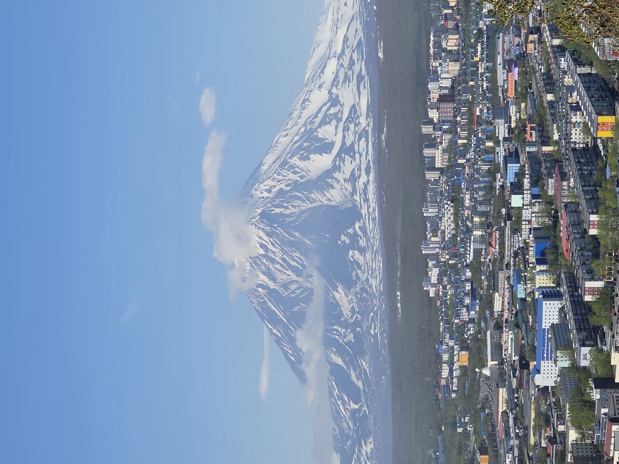 A vertical, high-angle, medium shot shows a snow-capped mountain with a plume of white smoke or cloud issuing from its peak, towering behind a dense, colorful cityscape under a clear blue sky. The mountain, likely a volcano, has a conical shape and is covered in snow on its upper slopes, with dark patches of rock or vegetation visible on its lower sections. A wispy cloud stretches horizontally across the upper portion of the mountain's peak.
Below the mountain, the city sprawls across the landscape, a mosaic of buildings with varied rooflines and facades in shades of blue, gray, white, yellow, and red. The structures range from low-rise apartment blocks to taller office buildings. Trees and greenery are interspersed throughout the urban area, particularly in the foreground and mid-ground. Roads and infrastructure are visible, connecting different parts of the city. The scene is bathed in bright daylight, casting subtle shadows. No individuals are visible in the image. The overall impression is one of a vibrant urban center set against a dramatic natural backdrop.