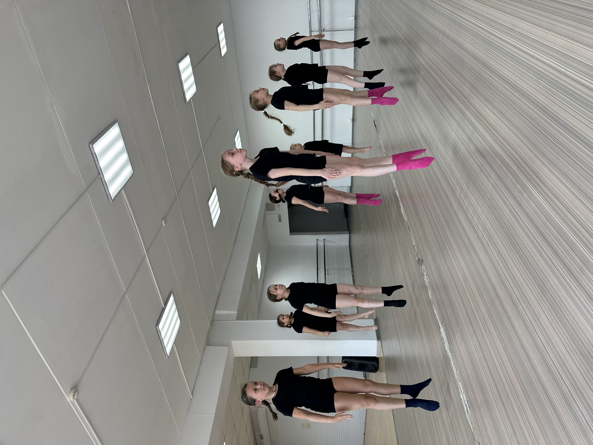 The image shows a dance studio in Votkinsk, Russia. A line of young girls wearing black leotards and socks of varying colors (pink, black, blue) are positioned near a wall of mirrors. They appear to be practicing a dance routine. In the background are ballet barres, standard in such studios. The room features a tiled floor and a ceiling with square fluorescent lights.