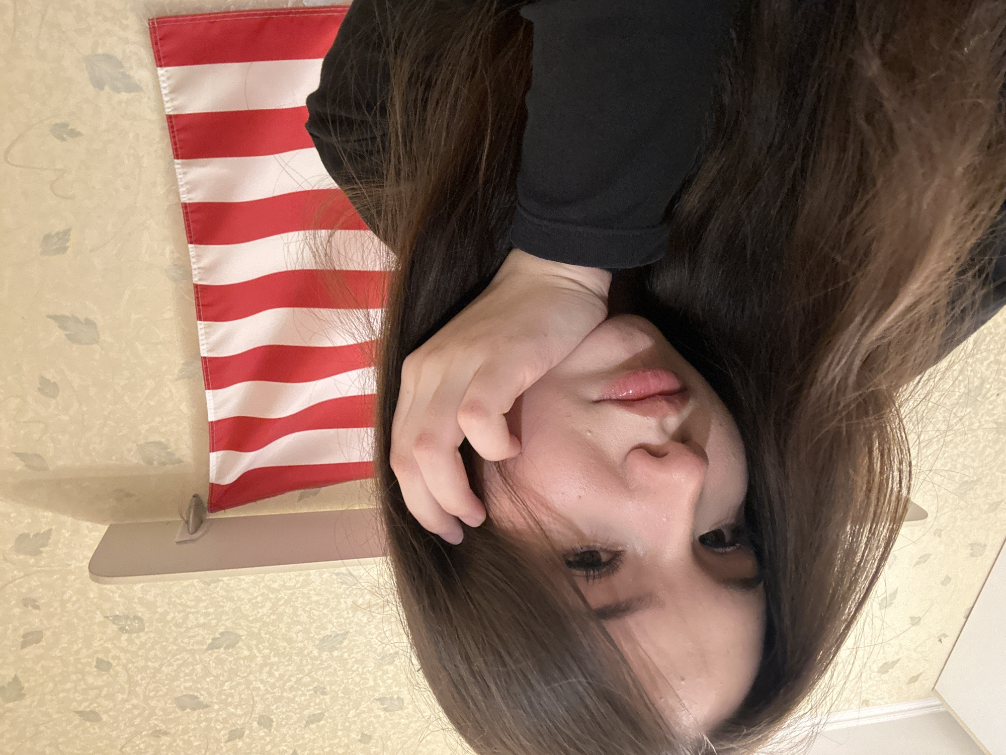 The image depicts a young female in an indoor setting, presumably a room within the city of Naberezhnye Chelny, Russia. The subject, a female with long brown hair, is positioned in the foreground, facing slightly towards the right side of the frame with her head angled downwards. Her left hand is raised, with fingers partially visible and her palm supporting her chin and jawline. Her expression is serious or contemplative, with her dark eyes looking slightly to the right of the camera. She has visible eyelashes and full lips, slightly parted. She is wearing a black long-sleeved garment.
The background consists of a light-colored wall, likely covered with wallpaper featuring a repeating pattern of faint, light grey-green leaves. In the upper left corner, a white ceiling molding and a segment of a white wall or door frame are visible. On the upper right, a white shelf is mounted horizontally on the wall. Directly below this shelf, a small American flag with horizontal red and white stripes is hanging vertically from a small, circular, silver-colored hook attached to the wall. The flag is partially draped and shows approximately seven red and six white stripes, consistent with the flag of the United States of America.
