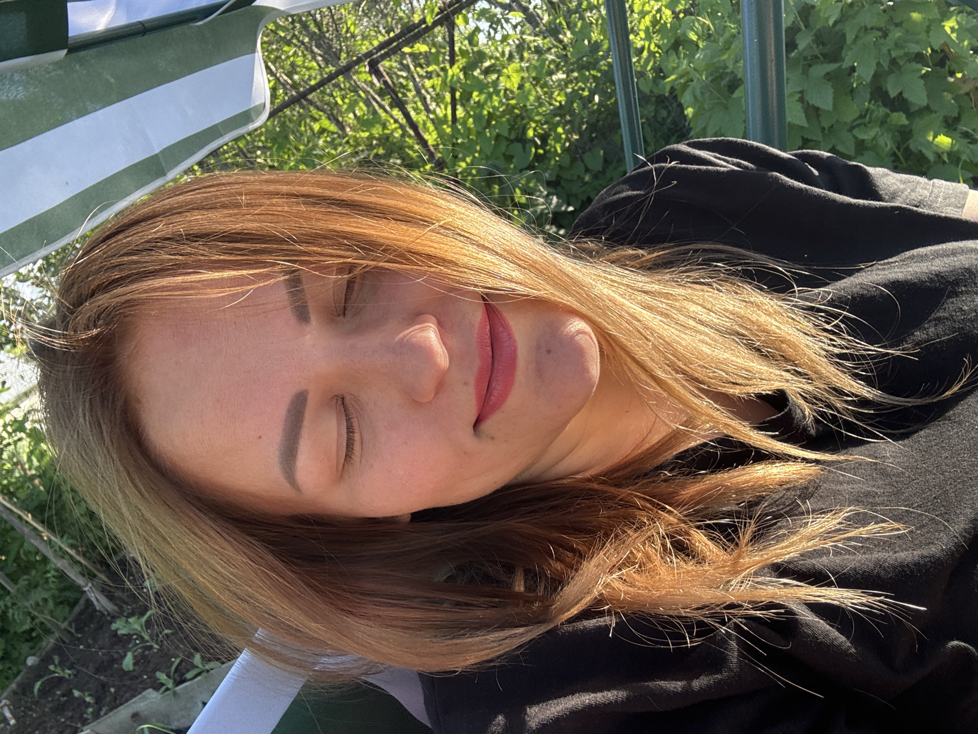 The image is a close-up, eye-level, outdoor shot of a woman in her mid-30s, with light brown hair, closed eyes, and wearing pink lipstick. She is wearing a black t-shirt. The background shows green foliage and a portion of a striped awning. The lighting suggests it is daytime, with sunlight illuminating the woman's face and hair. The scene appears to be a garden or park setting. The location is not specified within the image itself.