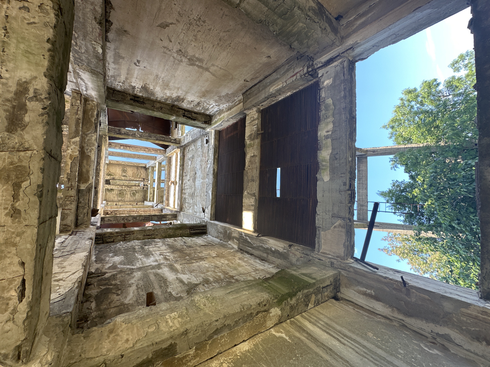 The image shows the interior of a derelict concrete structure, viewed from below, looking upwards towards the sky. The concrete is weathered and shows signs of decay, with cracks, peeling paint, and moss growth. Several floors or levels are visible, each with large openings that appear to have once held windows or ventilation grates, now partially covered by rusted metal sheeting.
In the foreground, the rough texture of the concrete walls and ceiling dominates. As the perspective moves upwards, the openings reveal glimpses of the sky and lush green trees. One opening at a lower level shows a metal ladder attached to the side of the concrete wall, suggesting access to higher parts of the structure.
The structure appears to be an abandoned industrial building or similar facility. The scene conveys a sense of decay and abandonment, with nature beginning to reclaim the man-made environment. The overall impression is one of a forgotten, possibly once functional, space left to the elements.
This structure is located in the city of Rosen, Bulgaria.
