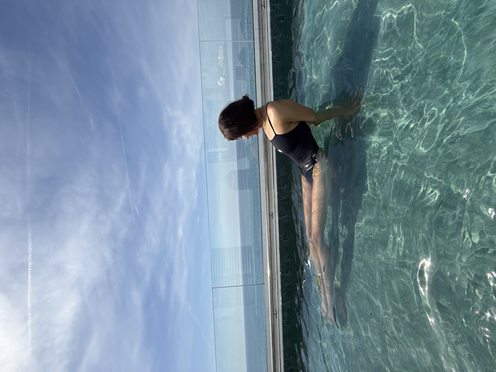 An adult female with short dark hair, wearing a black one-piece swimsuit with a white logo on the left chest, is partially submerged in an outdoor pool. She is positioned with her back to the viewer, looking towards her right (the viewer's left), with a slight smile visible. Her left arm is resting on the dark-colored edge of the pool, while her legs are extended forward under the clear, turquoise-hued water, which exhibits light reflections and ripples.
Beyond the pool, a transparent glass barrier separates the pool area from the expansive blue Mediterranean Sea, visible to the horizon. Through the glass, distorted reflections or views of distant architectural elements are subtly discernible. The sky above is a light blue with scattered, wispy white clouds and two distinct airplane contrails. The scene is illuminated by natural sunlight, indicating daytime in Calella, Spain.