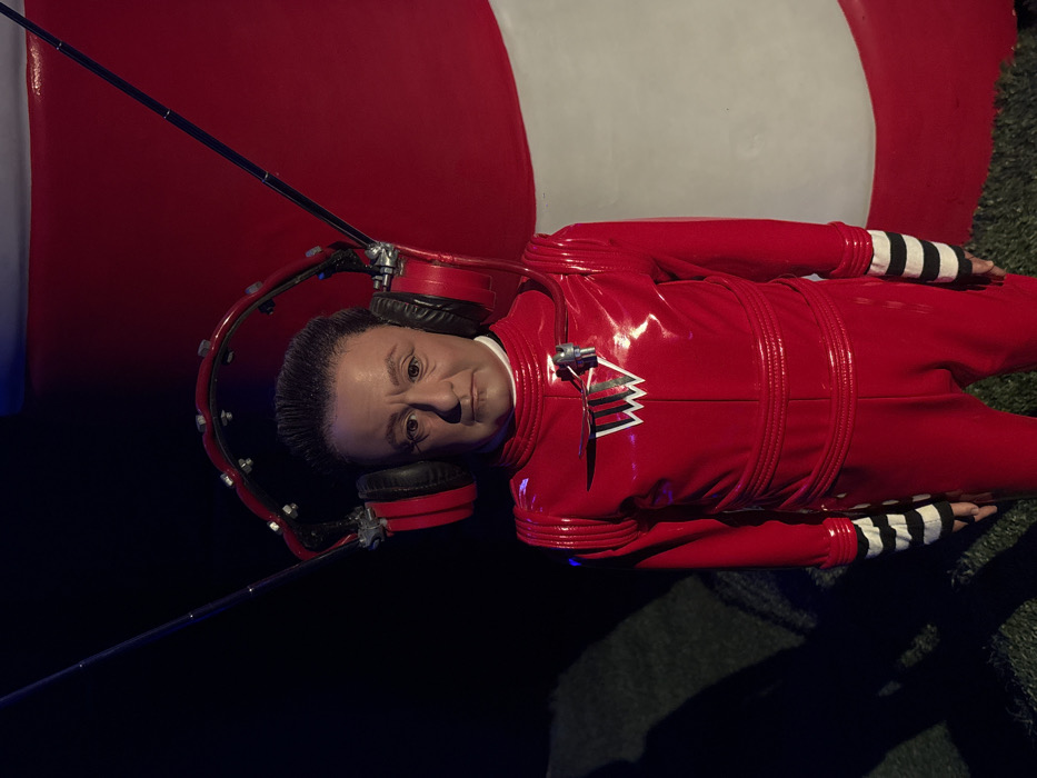 A wax figure depicting a character in a red jumpsuit with striped wrists, reclines in front of a red and white striped backdrop. The figure wears large red headphones connected by a metal band, featuring a long antenna. It has fair skin and dark hair, gazing directly at the viewer with a serious expression. The setting is dimly lit, with hints of artificial grass visible on the ground.