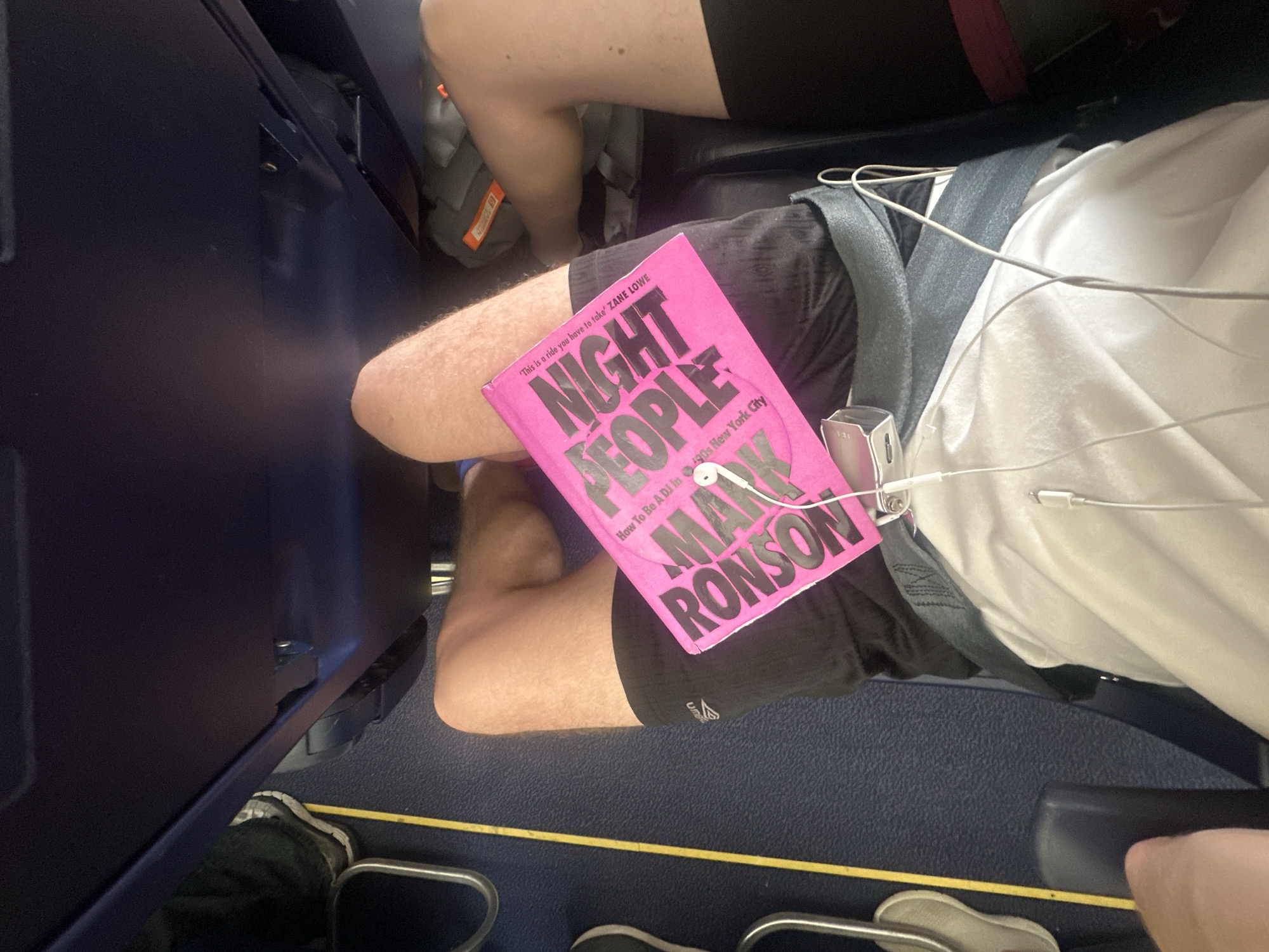 A passenger is sitting on an airplane, reading "Night People: How to Be a DJ In 1990s New York City" by Mark Ronson. The book is hot pink and resting on the person's lap. The passenger is wearing black shorts, a white shirt, and has headphones plugged into their ears. They are wearing a seatbelt. The interior of the airplane is visible, including parts of the seat, armrest, and floor. Another passenger's legs are visible in the background. Uttlesford, United Kingdom is not visible in the image.
