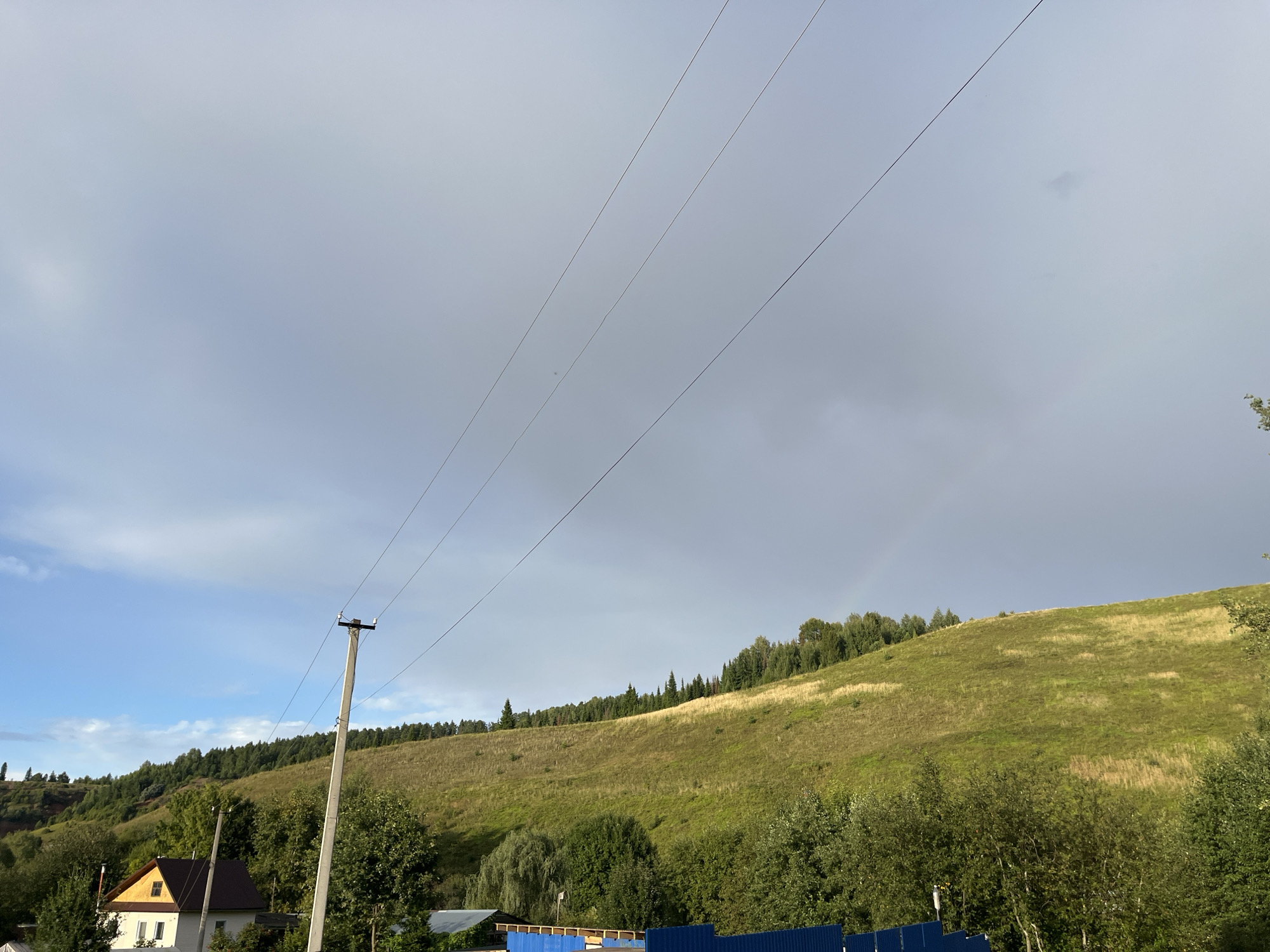 The image depicts a rural landscape in Olkhovka, Russia, dominated by a gently sloping, grassy hillside. A line of trees, primarily evergreens, crowns the hill, with a mix of deciduous trees and shrubs forming a dense border along its base. In the foreground, a small house with a brown roof and a white facade is visible to the left, partially obscured by trees. A wooden utility pole with visible wires stands prominently to the right of the house, stretching across the frame. The sky is overcast with patches of blue sky peeking through, and a faint rainbow is discernible in the upper right quadrant. The overall impression is one of serene, natural beauty.