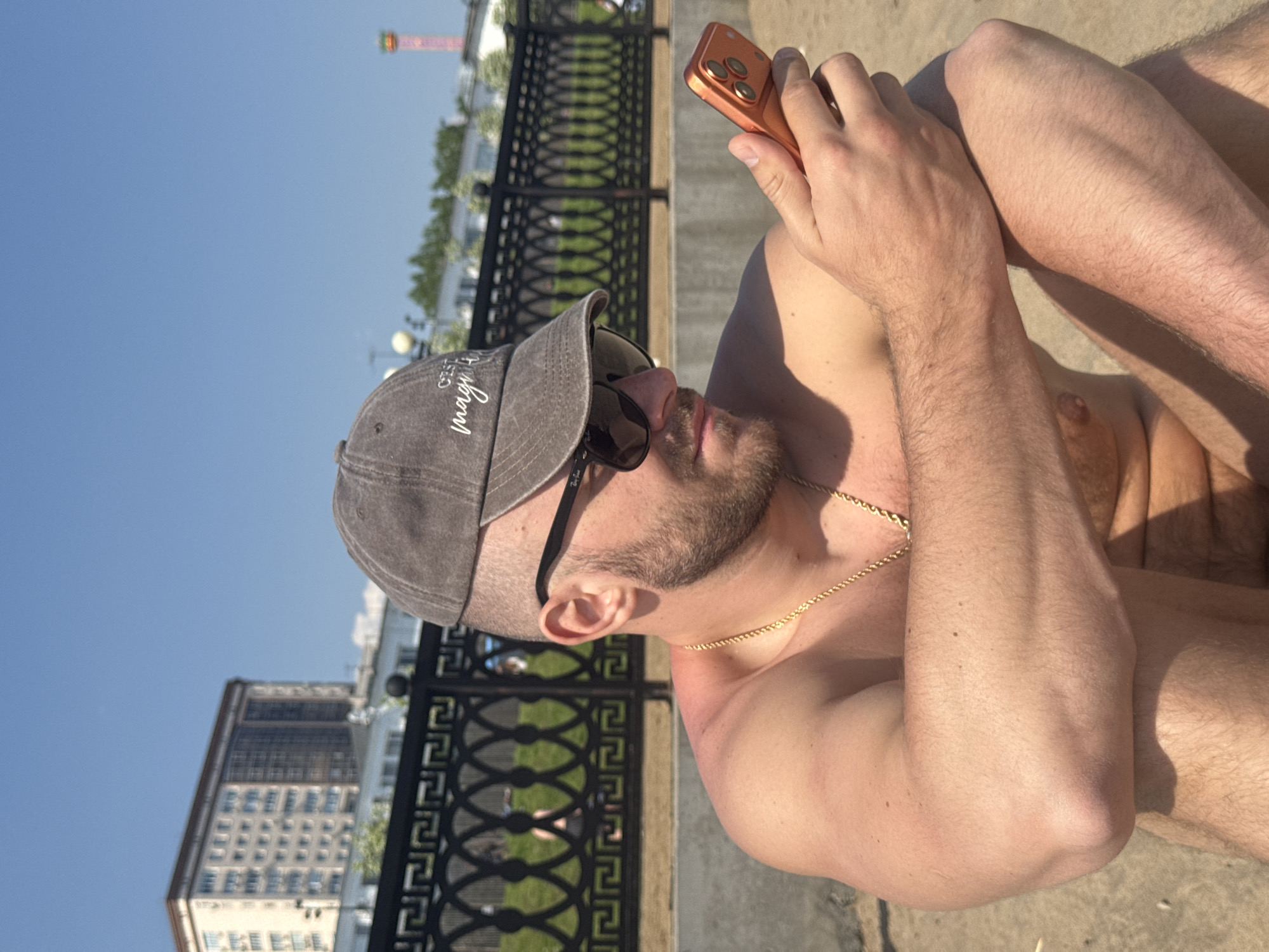 A man with a beard and a baseball cap, wearing sunglasses and a gold chain, is looking at an orange phone. He is sitting shirtless in front of an ornate black metal fence and a building. The scene is likely in Saratov, Russia, based on the skyline featuring a prominent tower similar to Saratov's observation tower.