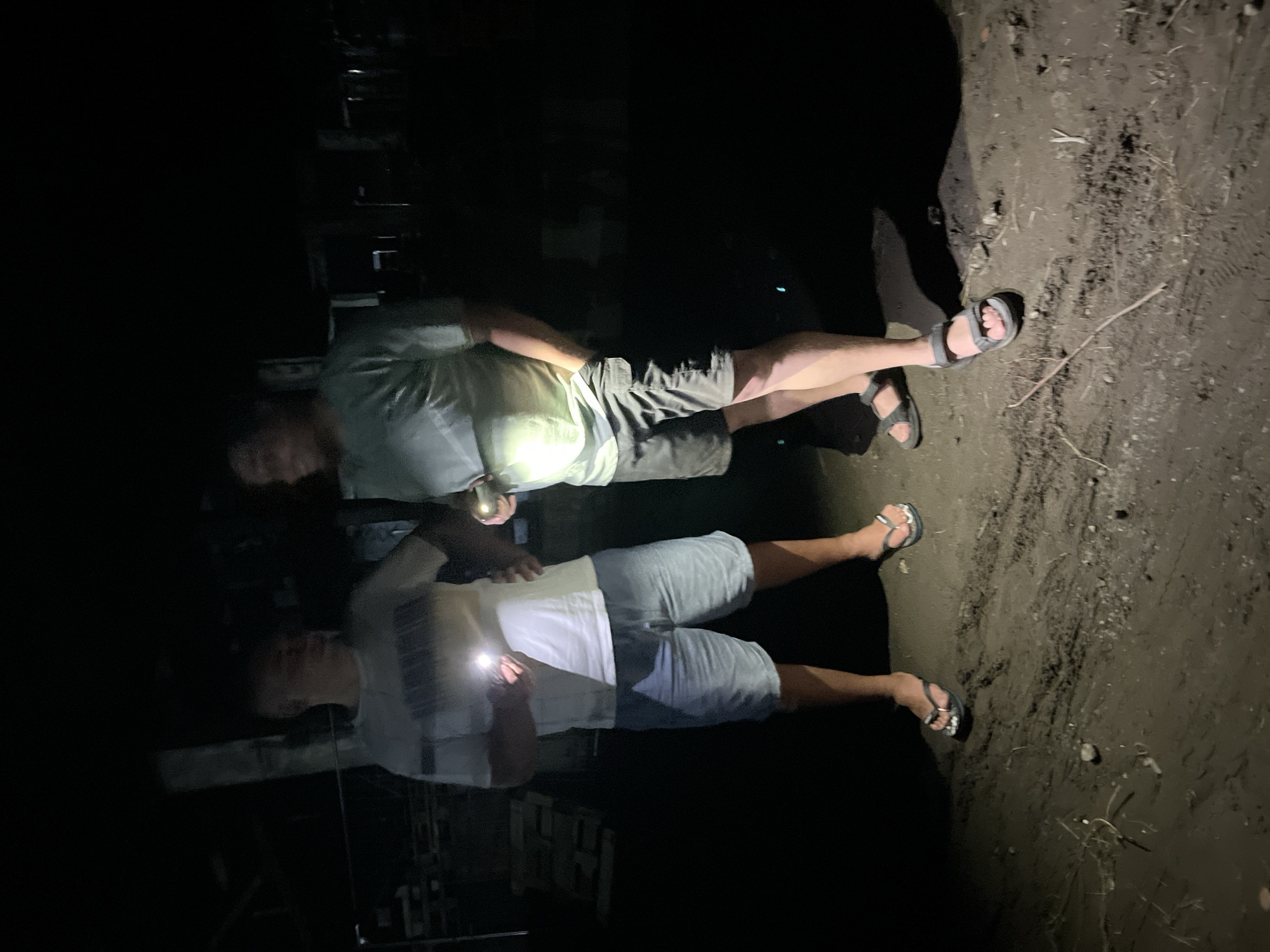 Two individuals stand outdoors in darkness, illuminated by flashlights. The individual on the left wears a white t-shirt with horizontal stripes and light blue denim shorts. They hold a flashlight in their right hand, the beam directed forward. The individual on the right wears a light-colored t-shirt and dark shorts, with their left hand in their pocket and their right hand holding a flashlight. The ground beneath them appears to be dirt and mud, with scattered debris. The background is largely obscured by darkness, but suggests an outdoor environment, possibly a rural area or a less developed part of the city of Poti, Georgia.