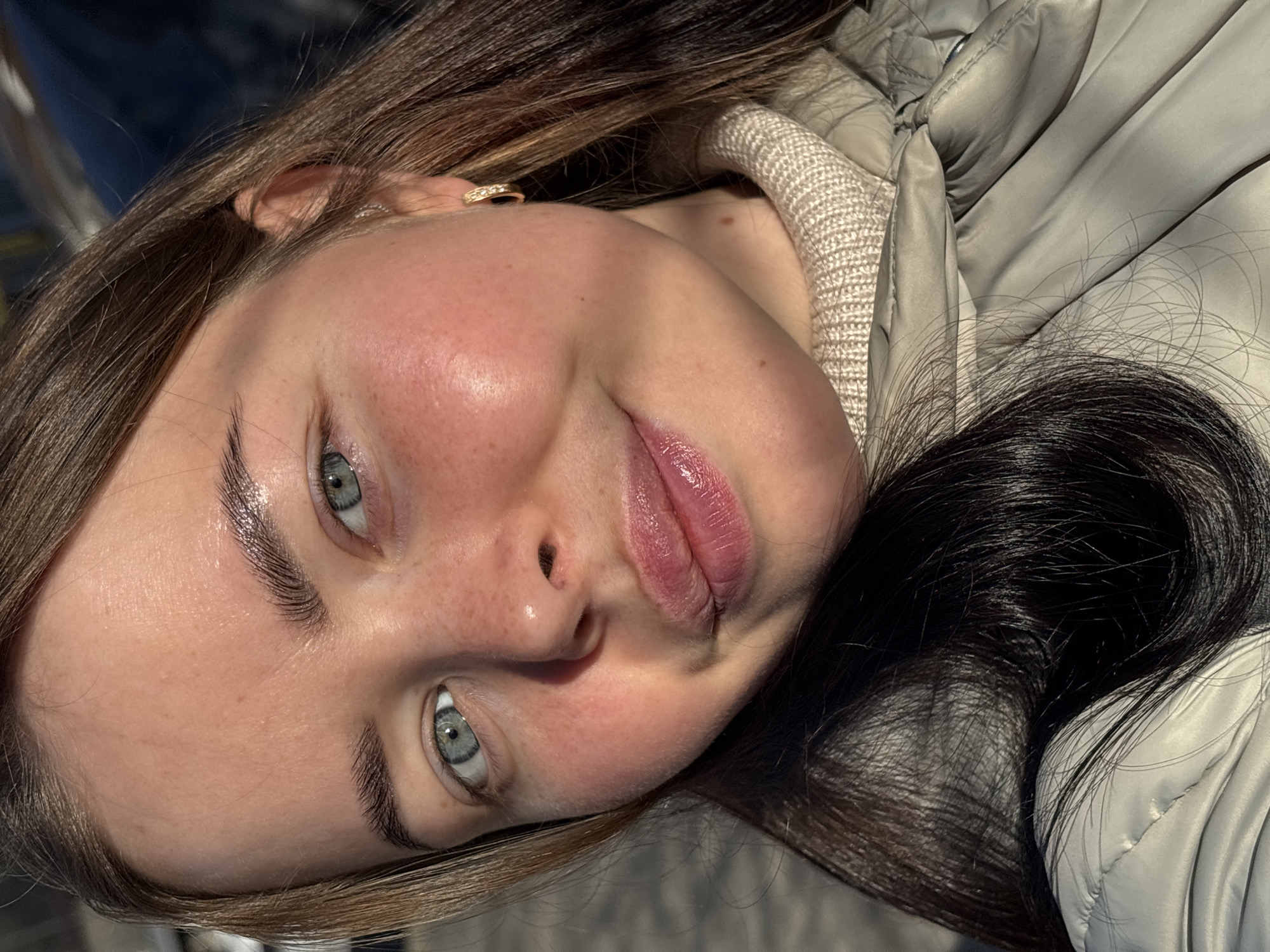 A close-up, slightly rotated portrait of a young woman with fair skin, gray-blue eyes, and full lips coated with pink lip gloss. She has light brown hair with dark brown streaks, visible near her face and on top of her head. She is wearing a white knit turtleneck sweater and a silver-gray puffer jacket. A gold ring is visible in her left ear. The background and specific location in Izhevsk, Russia are not discernible from the information available.