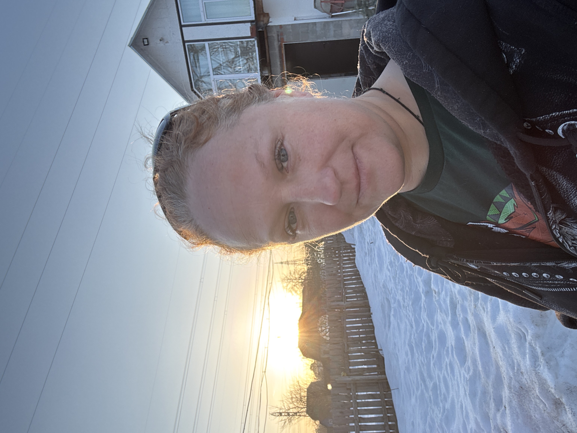 A selfie captures a fair-skinned woman with blonde hair and blue eyes in Chaikovsky, Russia. She wears a dark t-shirt and hooded jacket. The background shows a wooden fence, snow-covered ground, overhead power lines, a building with a window, and the setting sun.