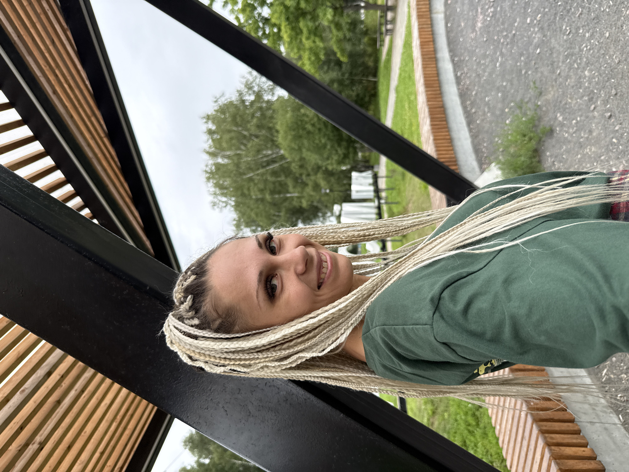 In Rusanovo, Russia, a woman with blonde braids stands under a wooden and black metal structure. She is wearing a green hooded sweatshirt. The background shows trees, grass, and benches.