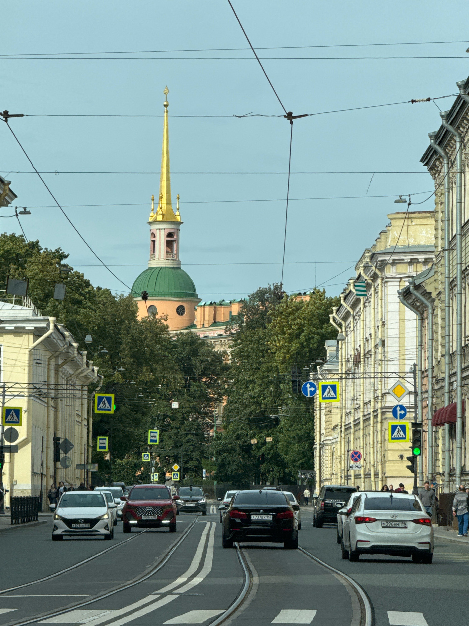 The image captures a view down a city street in Saint Petersburg, Russia, with a prominent golden spire of a building visible in the distance. The street is lined with multi-story buildings and trees. Several cars are driving on the road, which has tram tracks embedded in it. Pedestrians can be seen on the sidewalks. Traffic signals and pedestrian crossing signs are present. The sky is clear and light blue.