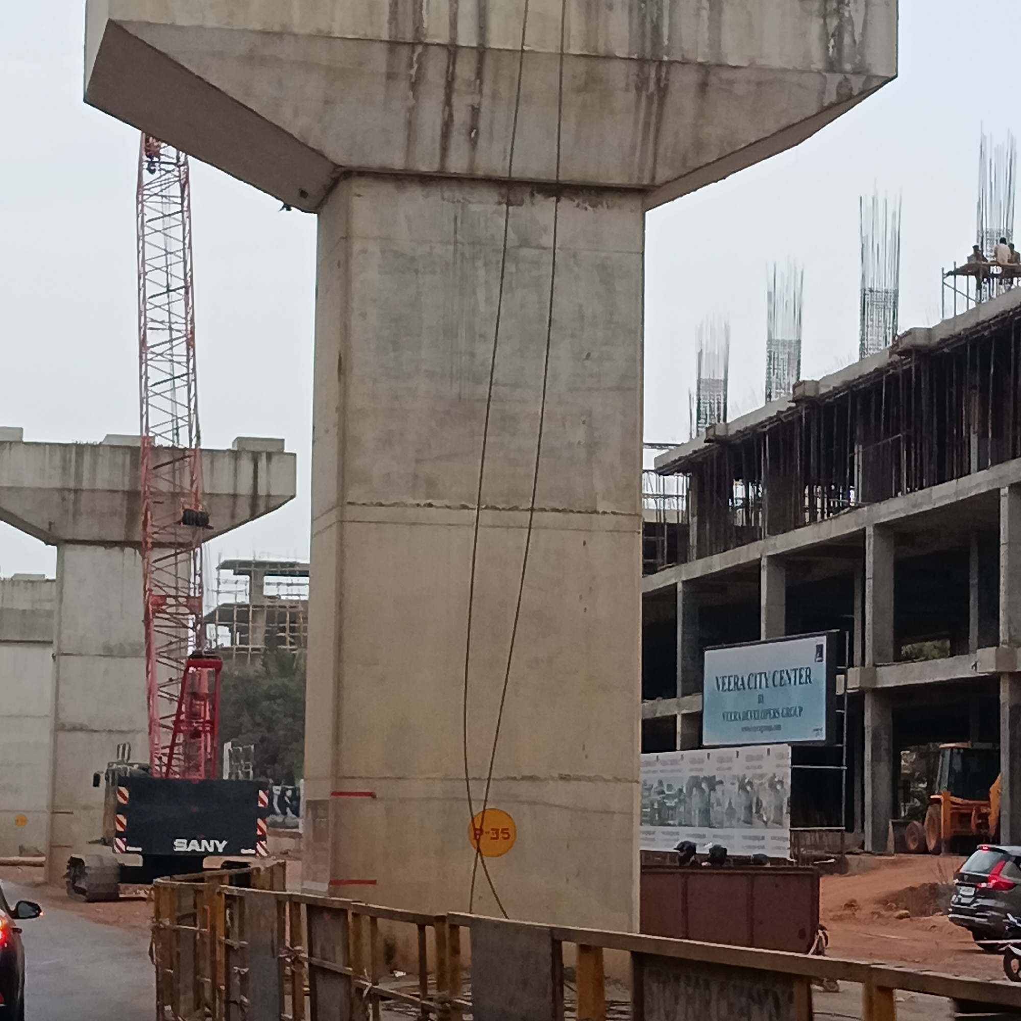 The image depicts an active construction site, likely for an elevated road or bridge project, situated in Sangolda, India. A large, gray concrete support pillar dominates the foreground. A red crane and another, similar pillar stand behind it. To the right, a partially constructed multi-story building, identified by a sign as the "VEERA CITY CENTER," is visible. Construction equipment like a backhoe and a SANY-branded tracked vehicle are present at the site. A car is partially visible in the lower left corner of the frame, and another vehicle can be seen near the VEERA CITY CENTER building. The scene suggests ongoing urban development in the area.