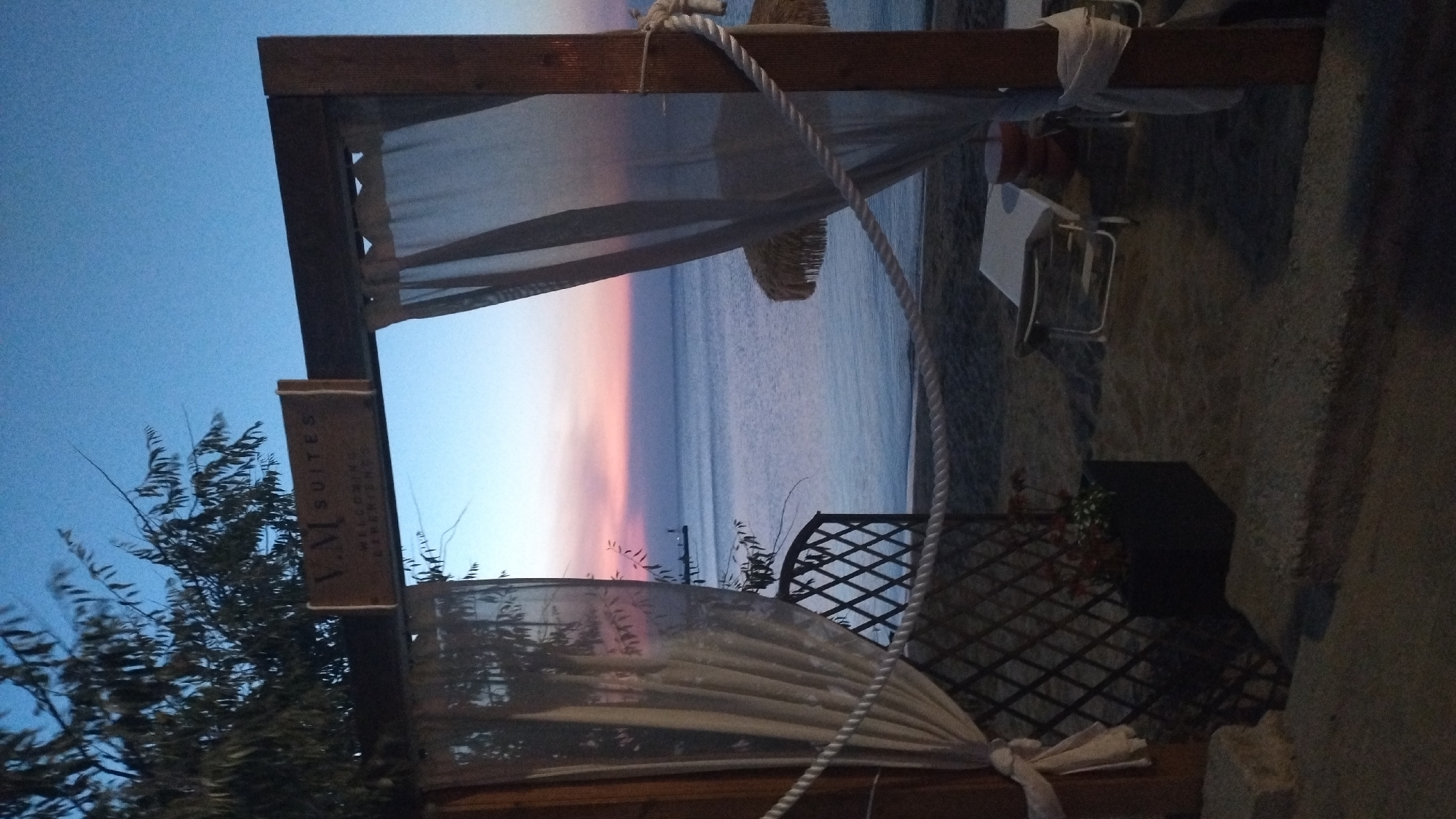 The image depicts an outdoor scene at dusk, likely at a beach resort in Neoi Epivates, Greece. A wooden archway draped with sheer white curtains frames a view of the sea. A sign above the arch reads "V.M. SUITES" and below it, "Welcoming Experiences". The sea is calm with a pastel-colored sky, transitioning from blue to pink. On the sandy beach, there are lounge chairs and thatched umbrellas. A dark planter box with red flowers sits near a diamond-patterned fence on the left side of the frame. The overall atmosphere is tranquil and serene, suggesting the end of a summer day.