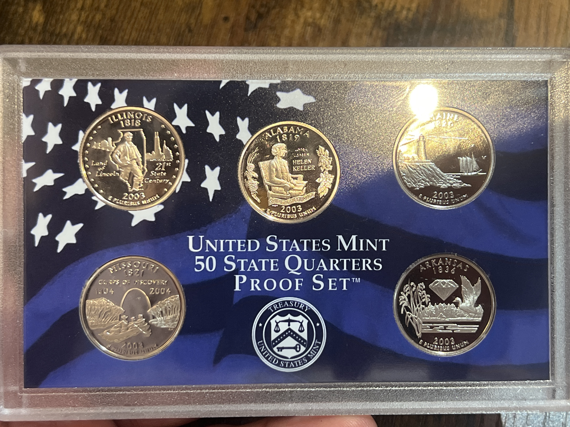 Here is a description of the image:
The image shows a "United States Mint 50 State Quarters Proof Set" in a protective case. The set includes five state quarters: Illinois, Alabama, Maine, Missouri, and Arkansas. Each quarter is designed with unique state-specific symbols and emblems. The case has a blue background with white stars, and the words "United States Mint 50 State Quarters Proof Set" are printed in white. There is a United States Mint logo with the word Treasury printed on it. The years when the states were founded are printed on the quarter. The words E Pluribus Unum are printed on the bottom of the quarter.