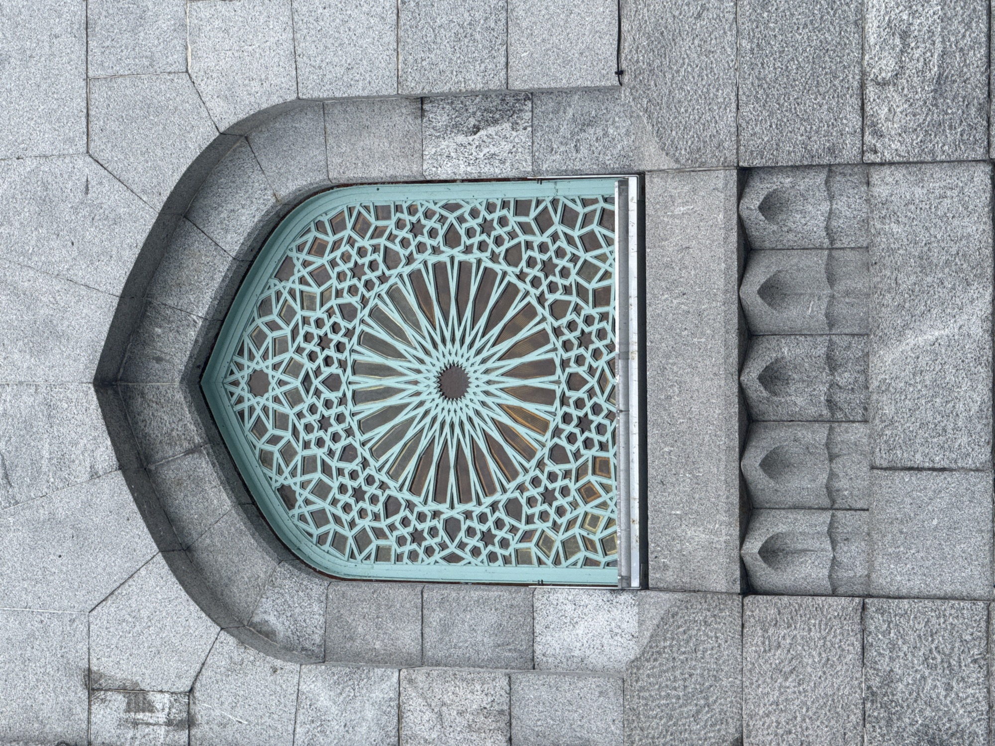 This overhead view from Saint Petersburg, Russia, presents a detailed architectural composition of stone and a decorative metal grate. The central feature is an elongated, pointed arch-shaped grate or window made of a patinated metal, exhibiting a distinct teal or verdigris color against a darker background. Its intricate geometric pattern includes a central starburst design from which numerous lines radiate outwards, forming a complex tessellation of stars, hexagons, and other polygonal shapes. This pattern is reminiscent of Islamic geometric art or ornate Art Deco detailing.
The ornate metalwork is embedded within a structure composed of light grey granite blocks. On the left side, the granite forms a smooth, sweeping curve that perfectly outlines the pointed arch of the decorative insert. The individual granite blocks in this curved section are precisely cut and fitted.
To the right of the central patterned element, the granite surface transitions into a series of vertically stacked, deeply recessed stone carvings. These appear as three-dimensional, sculpted forms, possibly functional steps or purely decorative fluting, adding depth and texture to the composition. The surrounding granite blocks display a rough, speckled finish, contrasting with the smooth edges of the metal frame.
The lighting is soft and diffuse, suggesting an overcast day or indirect light, which highlights the textures and dimensional qualities of both the stone and the metalwork. No activities or people are visible, and there is no discernible text in the image.