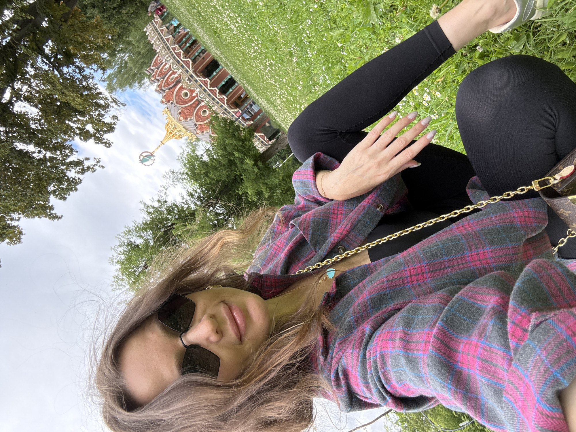 A woman wearing a pink and gray plaid shirt, black top, and black leggings is sitting on a grassy lawn dotted with white flowers. She is wearing sunglasses and a gold chain necklace with a turquoise pendant. In the background, a red and gold building with an ornate roof and a spire is visible, surrounded by trees. The sky is cloudy. The building in the background is the "Temple of the Buddha's Footprint" located in Moscow, Russia.