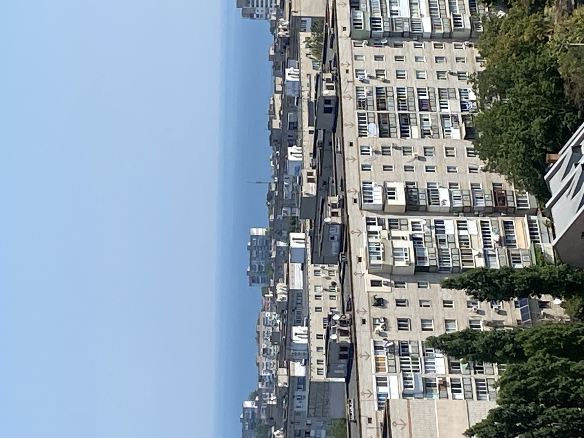 The image captures an aerial view of a dense urban landscape in Chornomorsk, Ukraine, with the Black Sea visible in the background. The foreground and mid-ground are dominated by numerous Soviet-era apartment buildings, characterized by their uniform concrete facades, repetitive window patterns, and numerous balconies. Many of these buildings feature external air conditioning units and satellite dishes. Anchor motifs are visible on the facade of the nearest building, suggesting a maritime connection. Tall, dark green coniferous trees are interspersed between and in front of some of the buildings, adding a touch of nature to the otherwise stark architectural setting. The sky is a clear, pale blue, indicating fair weather. No people are discernible in the image. The overall impression is one of a typical residential district in a coastal Ukrainian city.