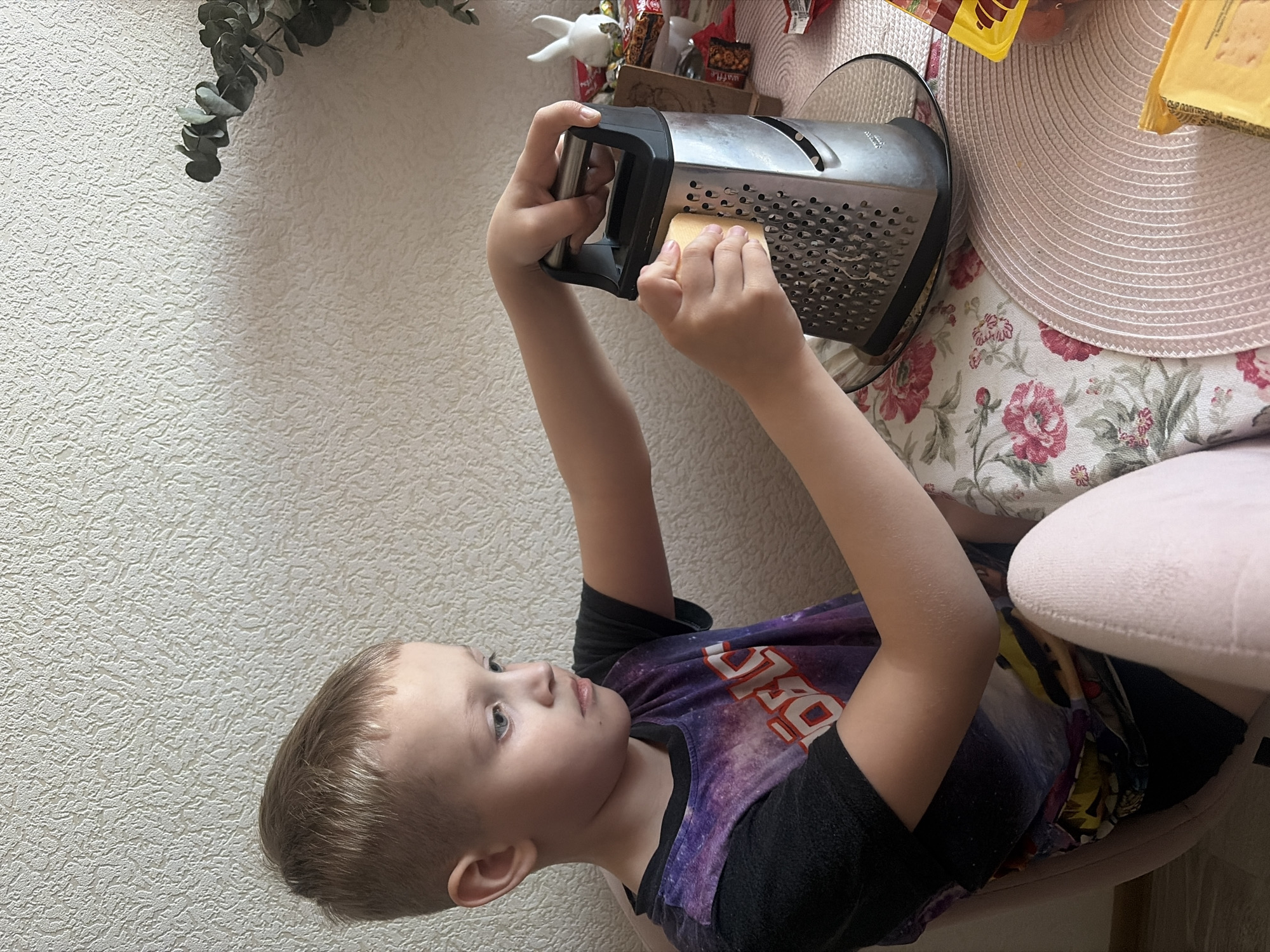 A young boy is grating cheese at a table. The boy has blond hair and is wearing a black and purple space-themed shirt. He is holding a block of cheese and a metal grater, appearing to be in the process of grating the cheese. The table is covered with a floral-patterned tablecloth and a pink placemat. Various items, including food packaging and a small figurine, are on the table. The background consists of a textured off-white wall and a sprig of greenery. The location is not discernable as being in Saint Petersburg.