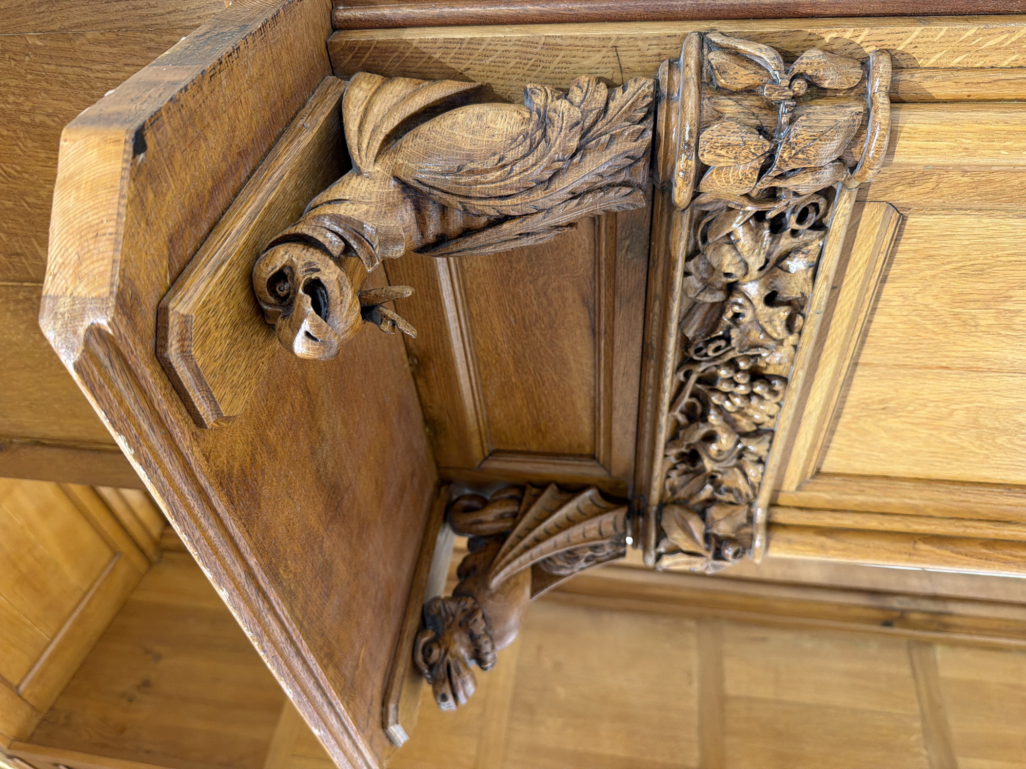 This is a close-up, eye-level, medium shot of intricately carved wooden architectural elements. The focal point is a wooden shelf supported by two corbels carved in the likeness of mythological creatures resembling dragons or griffins. The corbels have detailed wings, clawed feet, and expressive faces with open mouths and sharp teeth. Below the shelf, a decorative frieze is carved with foliage and clusters of grapes. The entire structure is made of a warm-toned wood, likely oak, with a polished finish that highlights the grain and carving details. The background consists of more wooden paneling. The lighting is even, casting subtle shadows that emphasize the depth of the carvings. The scene is static and devoid of any human presence. This image was captured in Palma de Mallorca, Spain, specifically within the interior of the Palau de l'Arquebisbe (Archbishop's Palace), a historical building showcasing rich architectural and decorative arts.