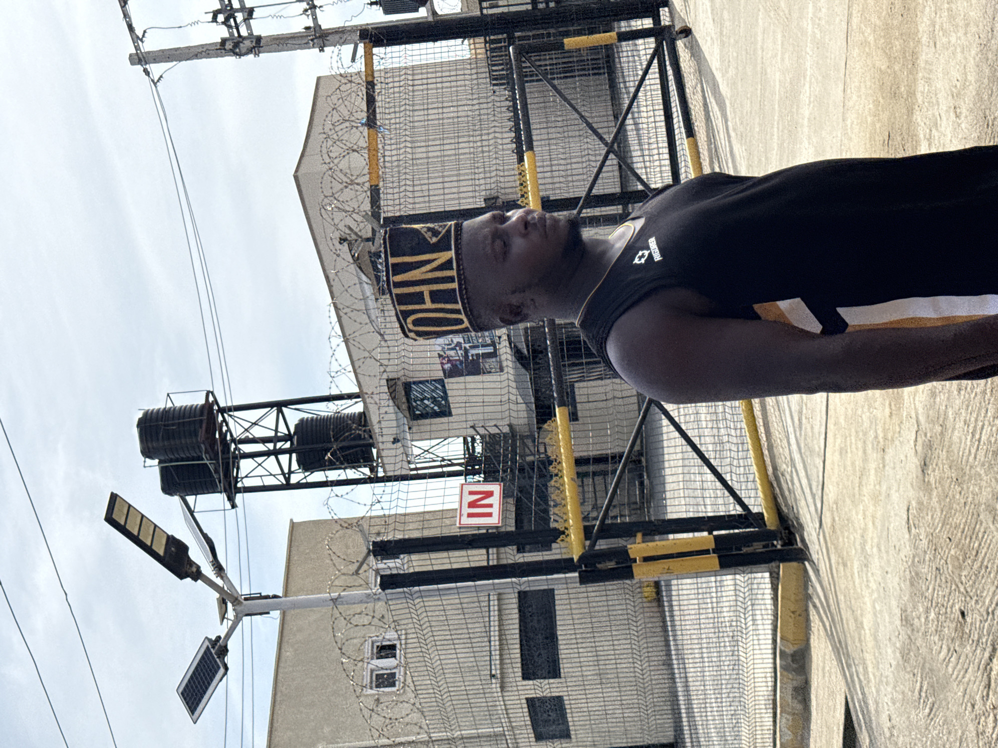 A medium, slightly angled shot depicts a person, presumably male, with dark skin, wearing a black tank top with white and gold accents, and a traditional black and gold cap with the name "JOHN" embroidered on it. He is standing in front of a tall metal gate with yellow and black posts. Behind the gate is a building with windows, and the gate itself is topped with barbed wire. A sign with the letters "IN" in red is visible on the gate. In the background, there is a pole with what appears to be solar panels and a street light attached, as well as an overhead power line. Above and behind the building, there are two black tanks held up by a metal frame. The scene appears to be taken in Ado, Nigeria, judging by the style of the clothing and the environment. The ground is concrete.