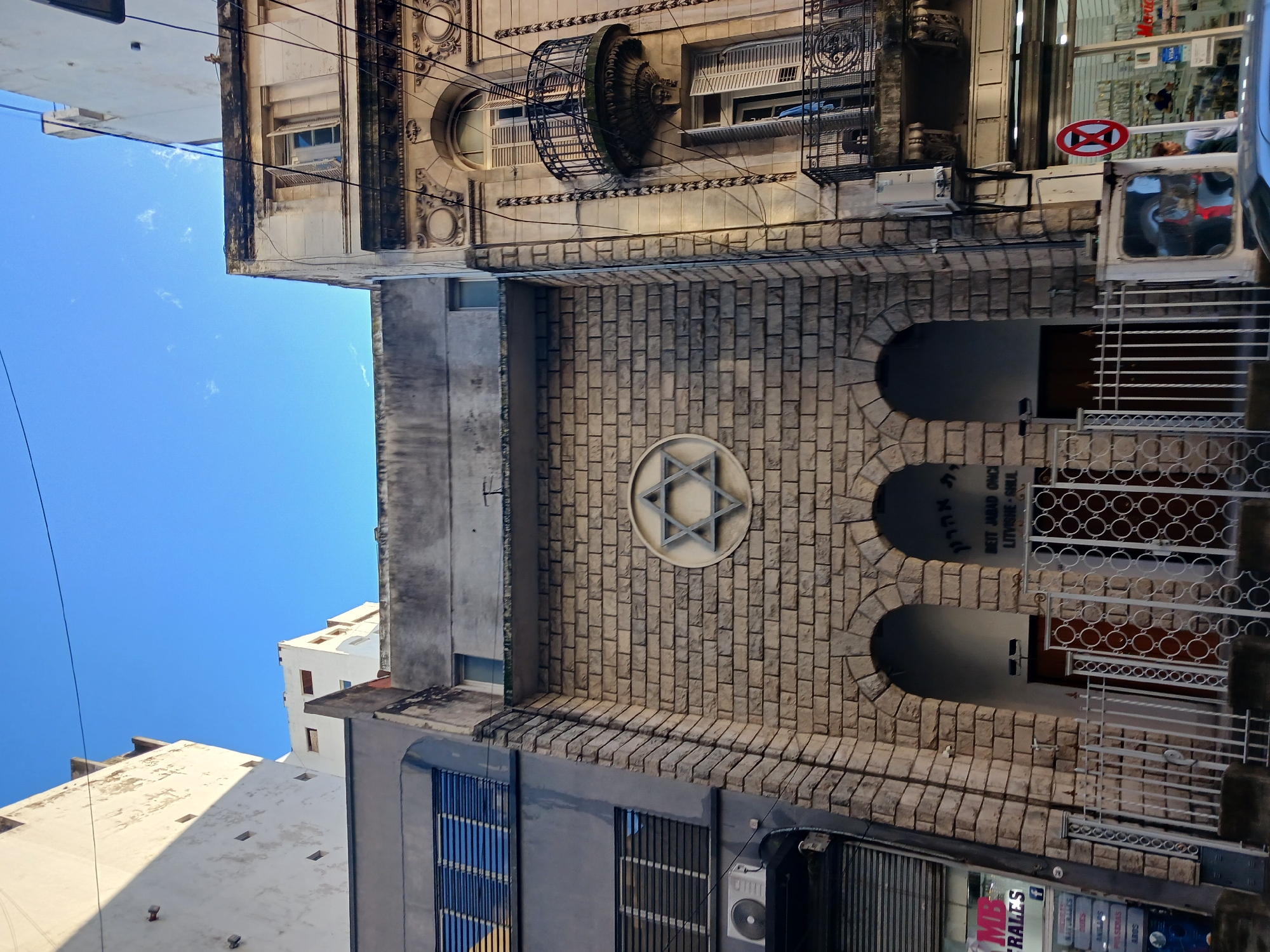 Here is a detailed description of the image:
**Scene:** An exterior shot of a building, specifically the Beit Jabad Once, a synagogue in Buenos Aires, Argentina.
**Architectural features:**
* The building is constructed of brick, particularly light-colored bricks.
* A prominent architectural feature is a large Star of David at the center of the brick facade.
* There are three arched entrances, each leading to a doorway within the synagogue.
**Surroundings:**
* The synagogue is situated in an urban environment.
* Adjacent buildings are visible on either side.
* Street-level businesses are indicated by signs and window displays.
* Other architectural details such as windows, balconies, and ornate facades are visible on the upper floors of adjacent buildings.
* An urban sky.