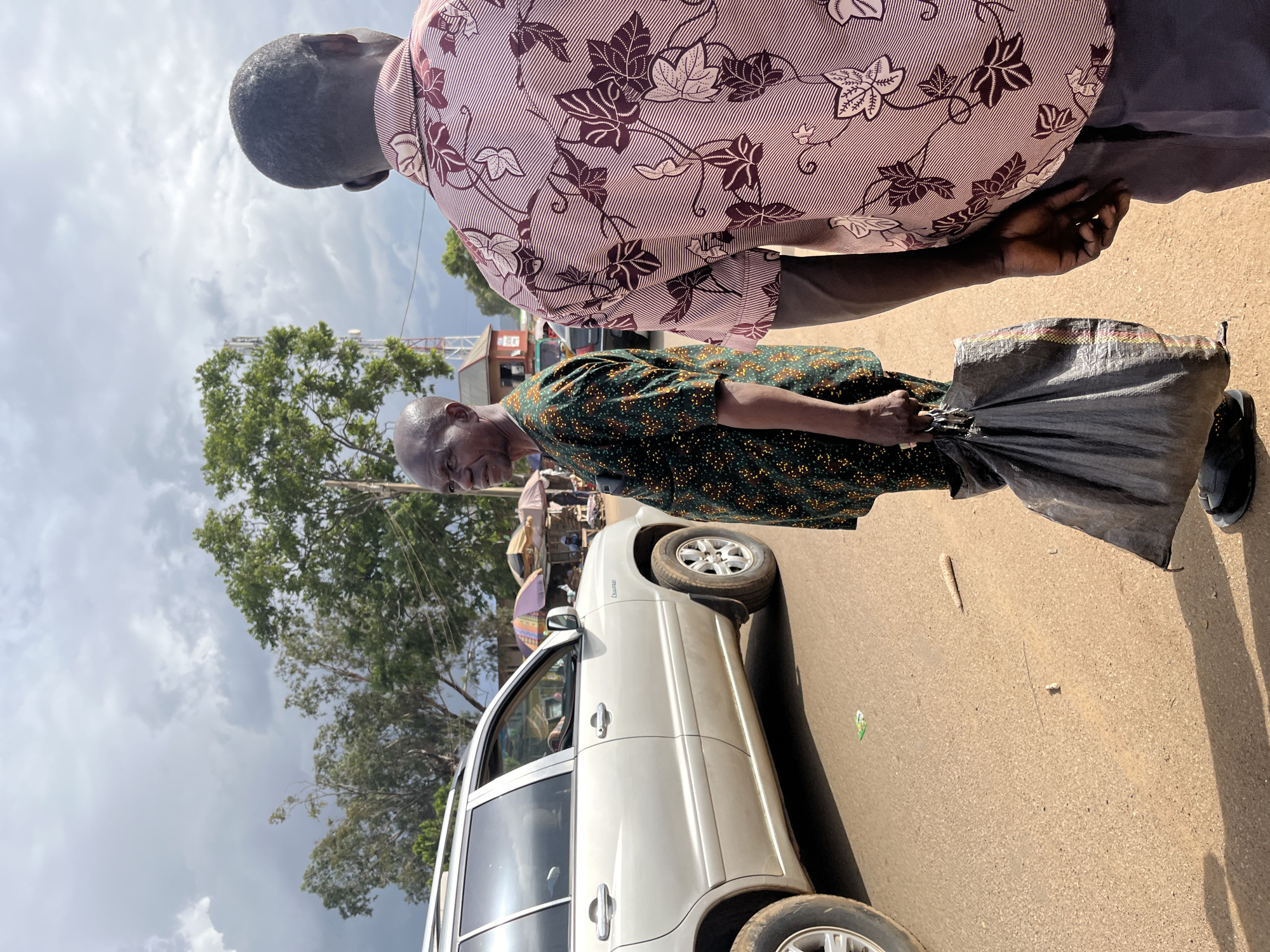 In Jos, Nigeria, two individuals are captured in a close-up, low-angle shot, suggesting a bustling street scene. The foreground is dominated by the back of a person wearing a pink and white patterned shirt with floral motifs and thin stripes. To their right, a second person, seen from the chest down, wears a green shirt adorned with small yellow dots. This individual is carrying a large, dark plastic bag in their right hand.
The ground appears to be a paved road, with a light-colored SUV parked on the left side of the frame. The SUV has its doors open, though the occupants are not clearly visible. In the background, a lush green tree stands tall against a cloudy sky, with hints of distant buildings and what might be market stalls. The lighting suggests it is daytime, possibly mid-morning or afternoon, with the sun casting shadows. The overall atmosphere seems ordinary and active, typical of a marketplace or roadside setting in Nigeria.