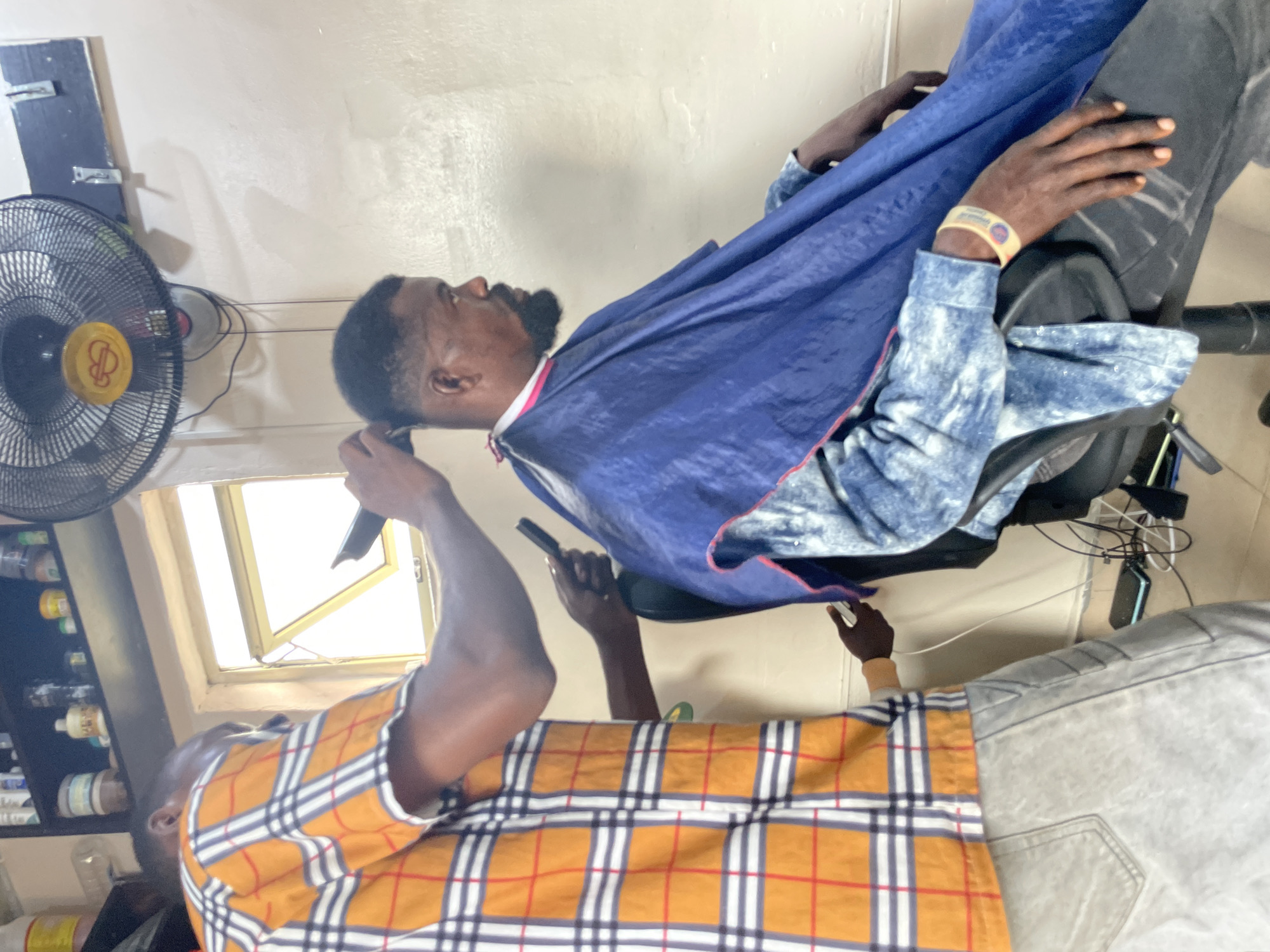Inside what appears to be a small barber shop in Ado, Nigeria, a barber with a brown and white plaid shirt is giving a haircut to a customer who is wearing a blue smock. The customer is seated in a black chair, facing the window. The barber is using clippers to trim the customer's hair. A fan is hanging on the wall. Various bottles are visible on a shelf.