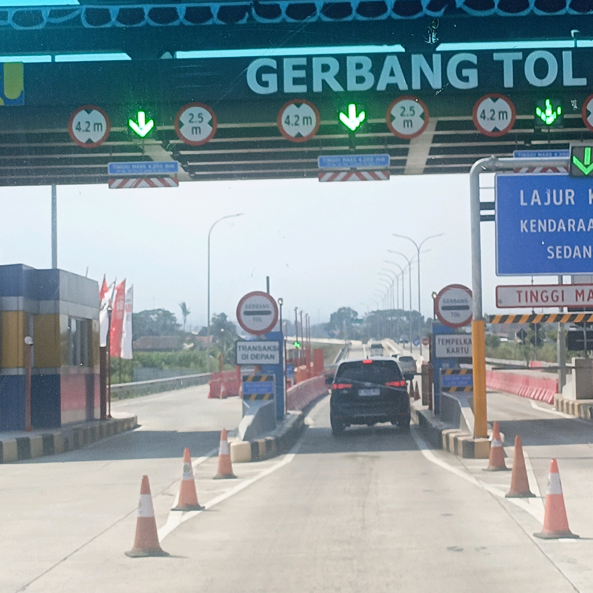 The image shows a toll gate on a highway in Dompyongan, Indonesia. The toll gate has overhead signs indicating "GERBANG TOL" (Toll Gate) and lane restrictions. Circular signs display height limits of 4.2m and 2.5m. Green downward arrows indicate active lanes. A blue sign reads "LAJUR KENDARAAN SEDAN" (Sedan Vehicle Lane), and another sign below it states "TINGGI MAKS" (Maximum Height). Red and white barriers are visible, along with orange traffic cones. Several cars are queuing at the toll gate, including a black SUV in the foreground. In the background, there are buildings, trees, and a bright, clear sky. Flags, including Indonesian flags, are displayed near a toll booth building on the left.