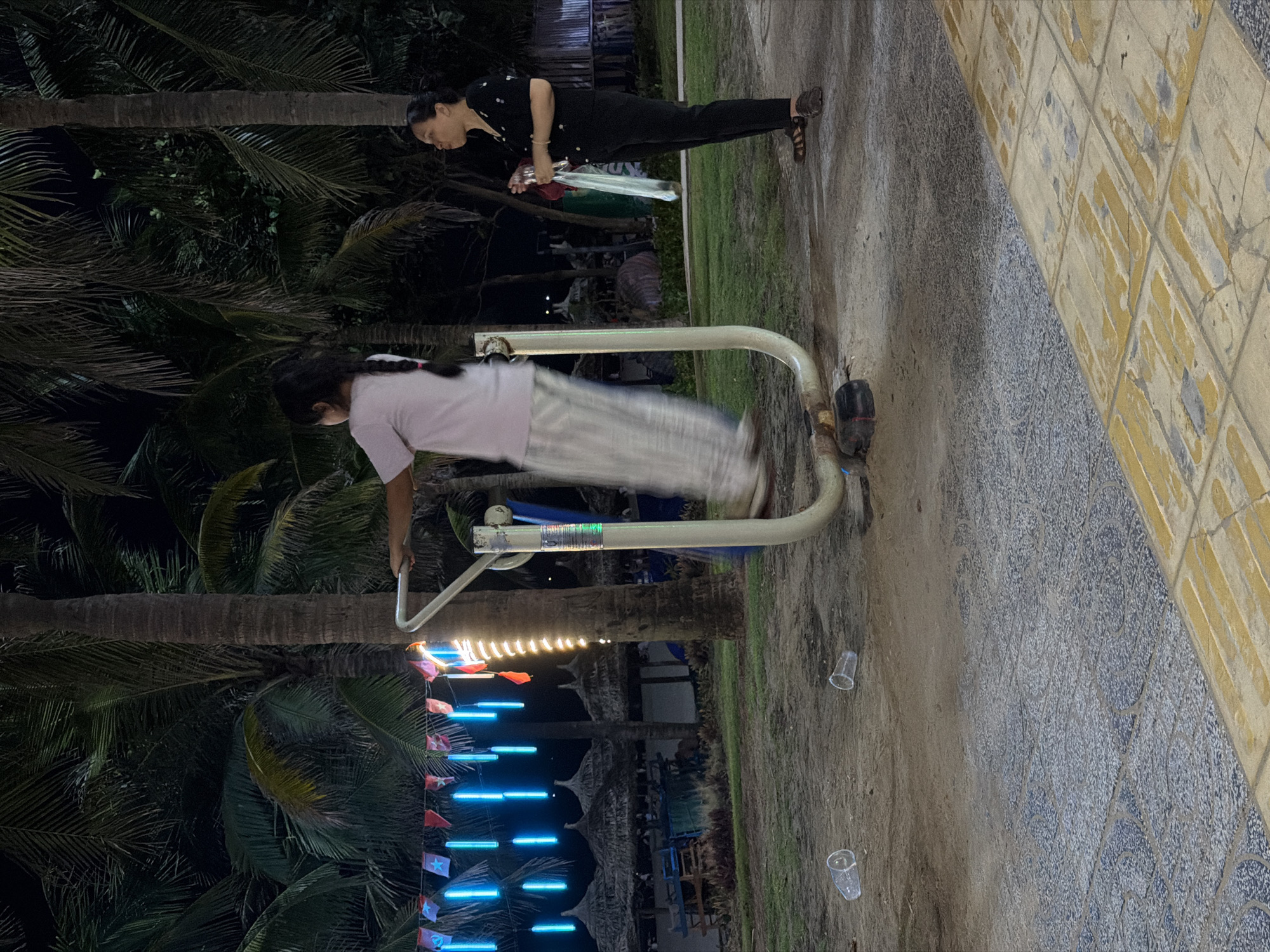 This media file shows two people in what appears to be an outdoor recreational area at night, likely in Đà Nẵng, Vietnam. One person, an adult female, is standing and holding a clear bottle and a green object, possibly a drink or a spray bottle. She is wearing dark clothing and sandals. The other person, a younger individual, is actively using an outdoor exercise machine, appearing to be in mid-motion. They are wearing a light-colored top and striped pants.
The setting includes palm trees, indicating a tropical or subtropical environment. The ground is a mix of sand and tiled paving. In the background, there are string lights illuminating some flags, creating a festive atmosphere. The overall lighting suggests it is nighttime, possibly late evening. The weather appears to be clear, as evidenced by the dry ground and clear visibility. The presence of plastic cups on the ground suggests a public area where people might have been relaxing or socializing. The mood seems casual and relaxed, with the younger person engaged in physical activity and the adult observing or preparing something. No visible text is present in the image.