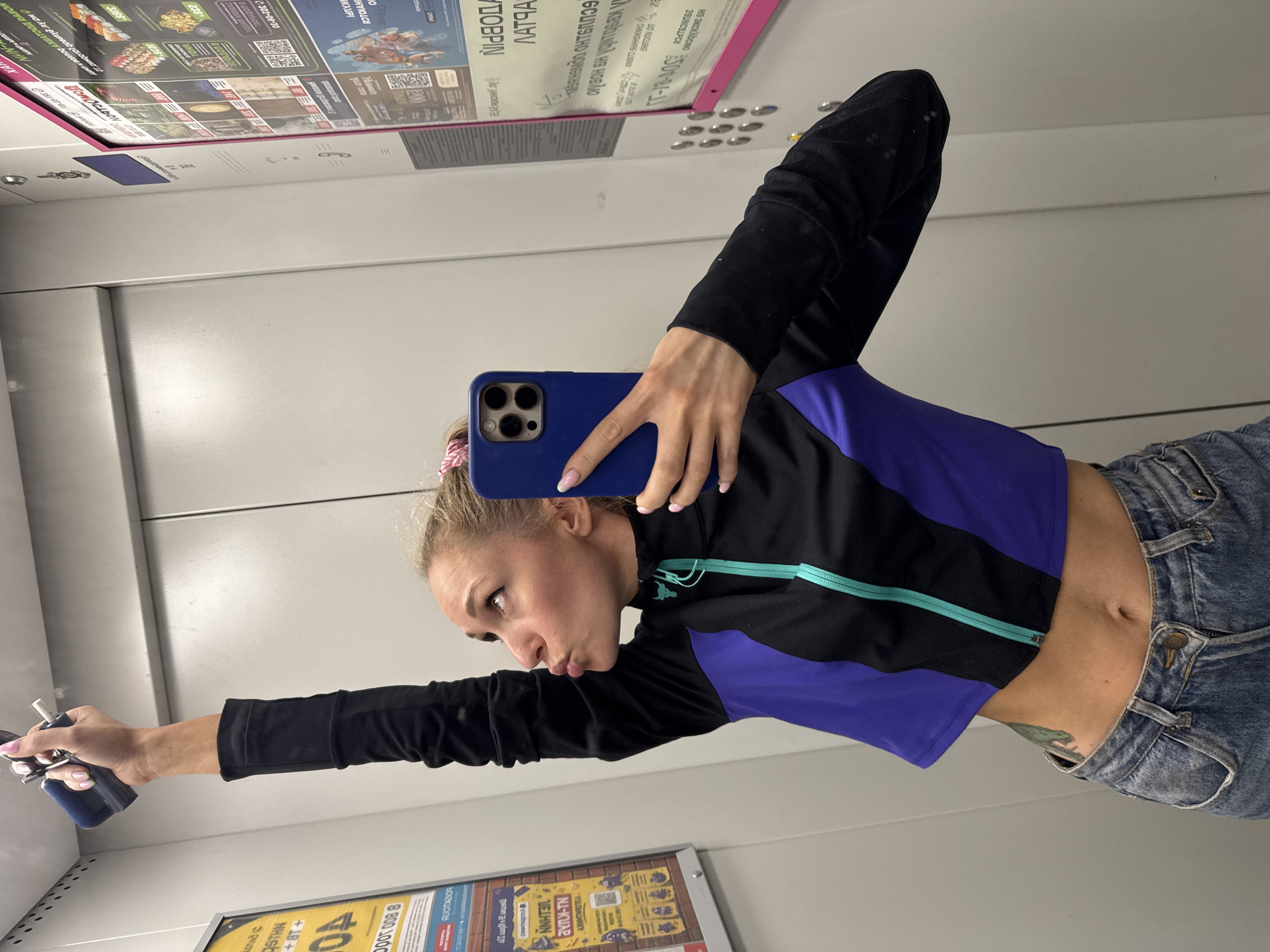 A blonde woman is taking a selfie in an elevator. She is wearing a cropped black and purple jacket with a teal zipper and blue jeans. Her left arm is raised, holding onto a handrail, and she is puckering her lips. She is also holding a blue smartphone in her right hand, taking the photo. The elevator walls are light gray, and there are posters on the right wall, one of which appears to be a menu for sushi. The elevator appears to be in the city of Perm, Russia, given the Cyrillic text on the posters. The overall impression is of a casual moment captured in an urban public space.