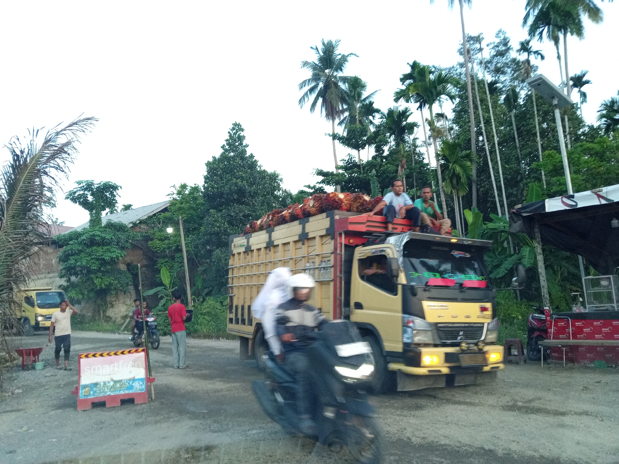 In Awegeutahpaya, Indonesia, a yellow truck loaded with palm oil bunches is parked on a dirt road. Two men are seated on top of the truck's load, with one man wearing a blue shirt and the other a green shirt. Another man in a light-colored shirt and dark pants is standing near a red roadside barrier, while a person in a red shirt and grey pants is standing further away. A yellow truck is visible in the background to the left, and a motorcycle with two occupants, one wearing a white helmet, is moving in the foreground. The environment features numerous palm trees and dense green foliage.