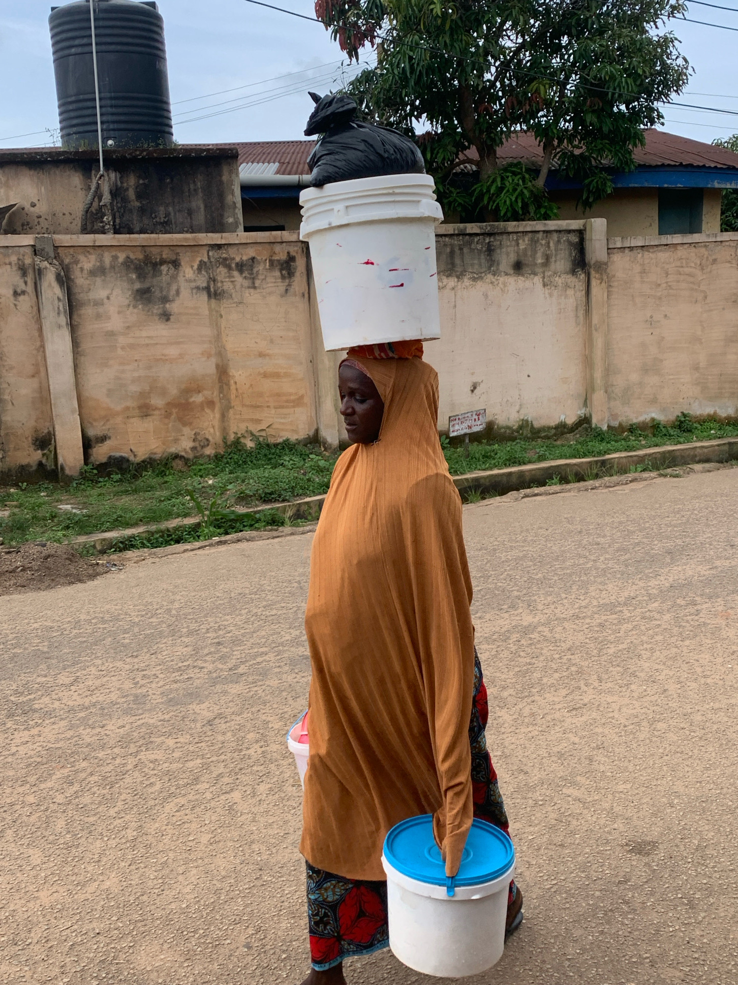 A person is walking on a paved road in Jos, Nigeria, carrying a large white bucket filled with a black bag on their head. The person is wearing a rust-colored headscarf and garment, and a patterned skirt. They are also carrying a smaller white bucket with a blue lid in their right hand. In the background, there is a beige wall with green grass growing in front of it, and a red-roofed building behind the wall. A large black water tank is visible on top of a smaller wall to the left. The sky is overcast.