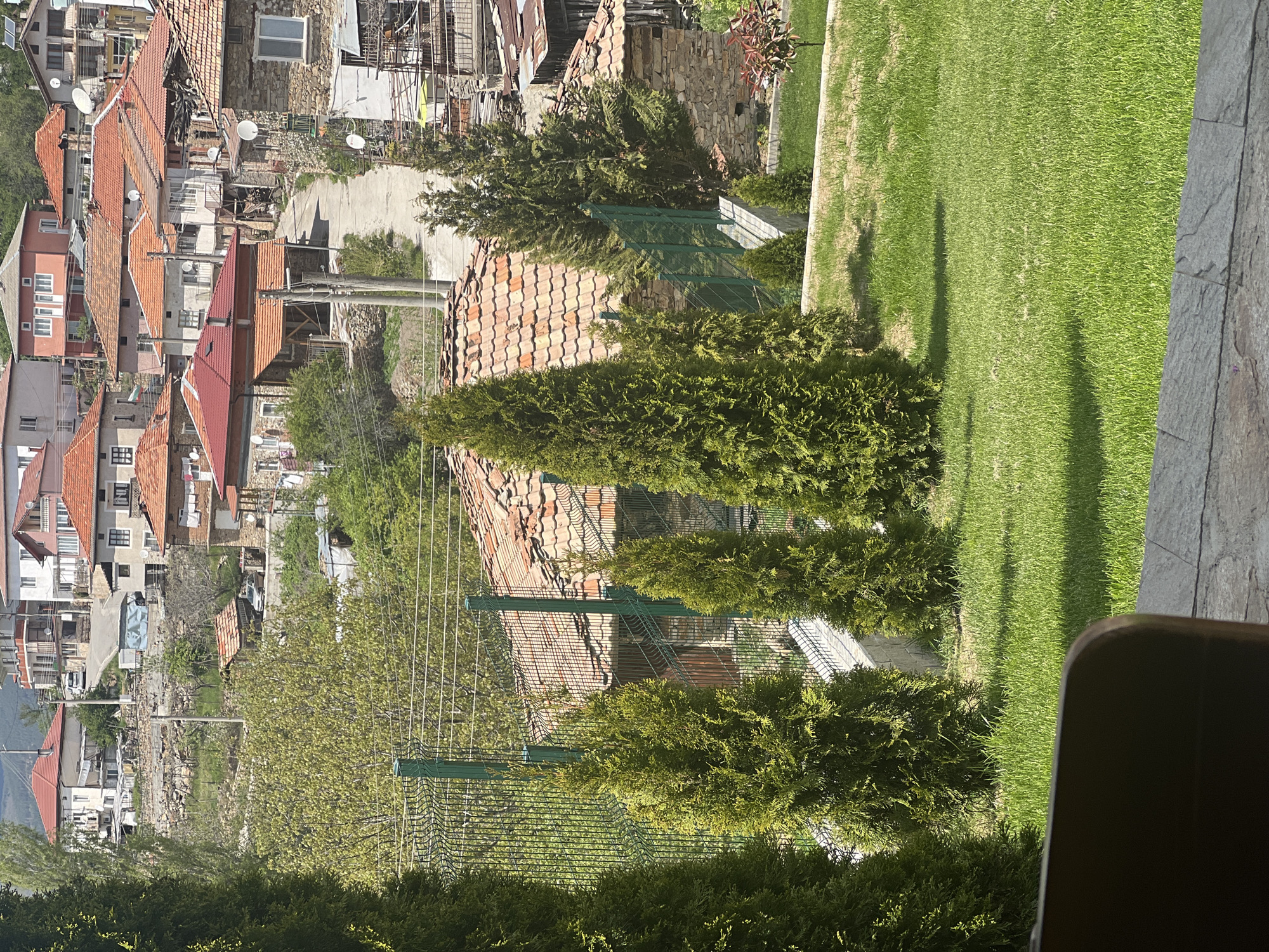 The image captures a panoramic view of a village, likely Kribul, Bulgaria, under bright, clear daylight. The perspective is from an elevated position, possibly a balcony or patio.
In the immediate foreground, a well-maintained, vibrant green lawn stretches across the lower portion of the frame, bordered on the right by a textured grey stone path or wall. Long shadows, presumably from trees, extend across the grass. A dark, indistinct object, possibly part of an armrest or furniture, is visible in the bottom right corner.
Beyond the lawn, a row of slender, dark green conifer trees (possibly thujas or cypresses) stands vertically, partially obscuring a green wire or chain-link fence. Behind these, the middle ground reveals a dense cluster of houses with characteristic red terracotta tiled roofs. The buildings exhibit a mix of older stone constructions and stucco-finished walls, some with visible windows and balconies. Numerous white satellite dishes are mounted on the rooftops. Overhead power lines crisscross the scene, supported by utility poles that snake through the village. A narrow, unpaved or concrete road winds between the houses. On one house in the mid-distance on the left, a small Bulgarian flag (horizontal white, green, red stripes) is visible.
The background shows more houses extending up a gentle slope or into the distance, blending with surrounding green foliage and hints of distant hills or mountains on the far left horizon.
The overall impression is one of a peaceful, traditional village setting bathed in strong sunlight, indicated by the distinct shadows and bright illumination. No people or specific activities are depicted; the scene is static and serene.
