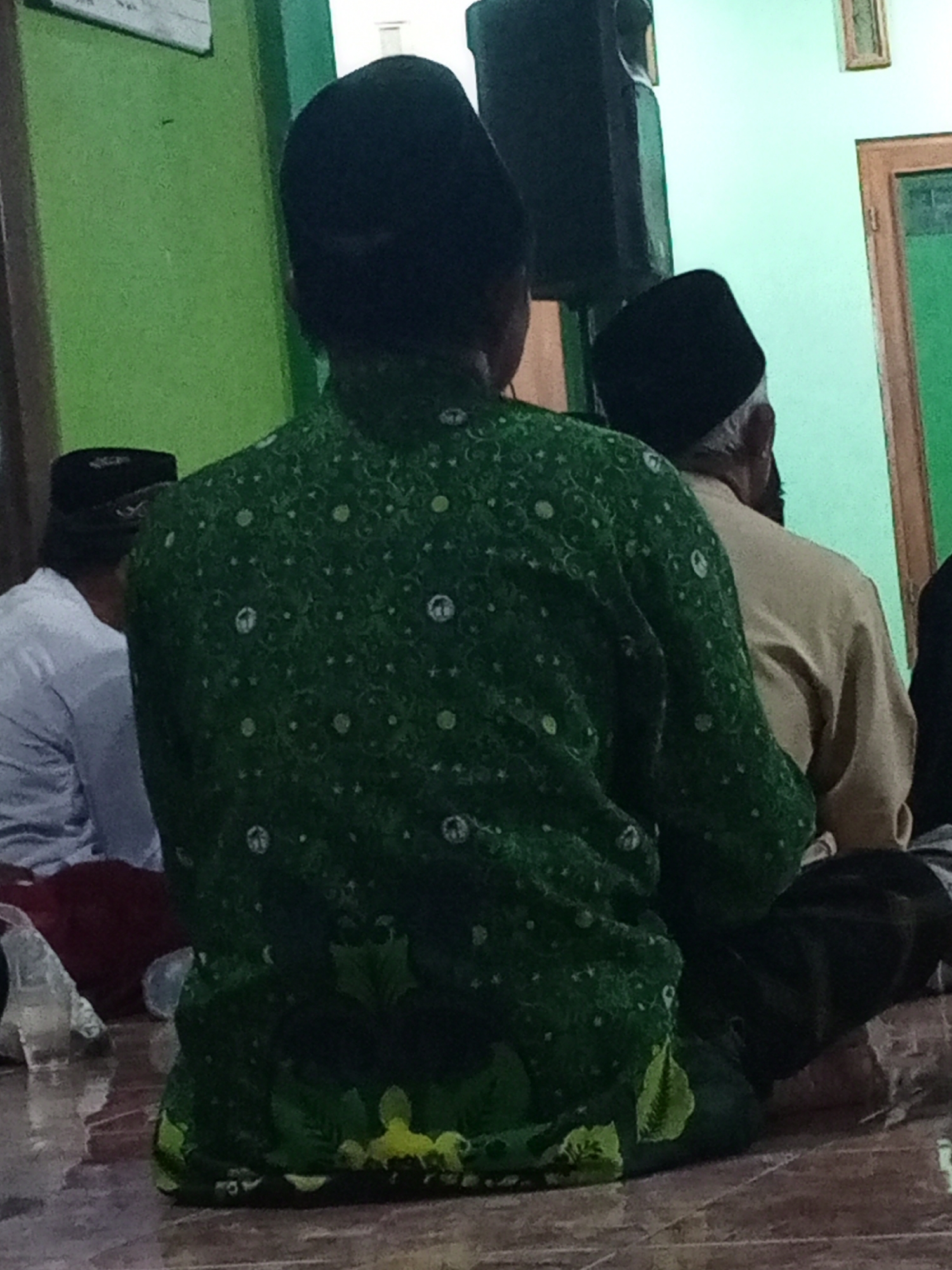 The image depicts three men indoors, seen from the back. The man in the foreground wears a black peci and a green long-sleeved shirt with a white circular pattern. He is seated on the floor, which is tiled in a wood-like pattern. To his left, another man also wears a black peci and a white long-sleeved shirt. Further to the right, a third man, older with graying hair, wears a black peci and a light beige long-sleeved shirt. He is also seated on the floor. Behind the seated men, a tall black speaker is visible against a green wall. A partially visible window with a wooden frame and green glass is present on the right side of the image. The overall environment appears to be a room, possibly a mosque or prayer hall, with plain green walls and tiled flooring. The men appear to be engaged in prayer or a religious gathering. The scene is captured indoors in Lemahbang, Indonesia.