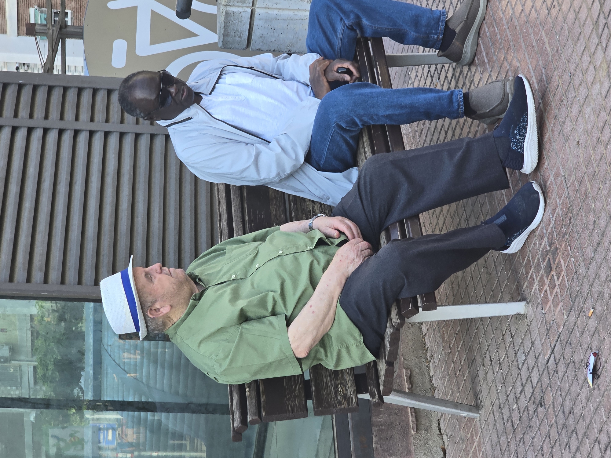 Two men are depicted sitting side-by-side on a wooden slatted bench in an urban outdoor setting in Cornellà de Llobregat, Spain. The image appears to be taken during the daytime under bright natural light.
On the left, an older man, likely white, is seated. He wears a white hat with a blue band, a long-sleeved light green collared shirt, and dark trousers. A watch is visible on his left wrist, and his hands are clasped in his lap. He looks forward or slightly upward with a calm expression. He is wearing dark sneakers with white soles.
On the right, a man, likely Black, is seated. He wears sunglasses, a light-colored (possibly white or light grey) hooded jacket, blue jeans, and dark, possibly slip-on shoes. His hands are clasped in his lap, and he also appears relaxed, looking forward.
Behind the men is a tall, corrugated metal or plastic wall structure. Above this wall, to the right, a large, light-colored sign with abstract dark symbols or partial letters (including what looks like an 'N' shape) is visible. To the left of the men, a large glass surface reflects trees, buildings, and the sky.
The ground in front of the bench is covered with reddish-brown pavement tiles featuring a diamond pattern. A cigarette butt and a small piece of paper or rubbish are visible on the pavement. Both men appear to be resting or waiting, contributing to a tranquil and casual atmosphere. No decipherable text is clearly visible on the background sign.