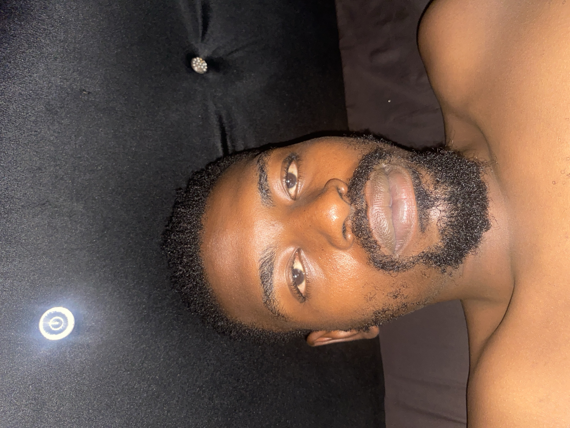 A close-up, sideways portrait captures an individual, likely male, with dark skin and a short, dark, well-groomed beard. The person's head is tilted, and they are looking directly into the camera with a neutral expression. Their left eye is prominent, with dark eyebrows and lashes, while the right side of their face is partially obscured. Short, dark, curly hair frames the face.
The individual is situated against a dark, possibly upholstered surface, which features a visible decorative, studded button, suggesting a tufted headboard or cushion. To the left, a faint, circular, glowing object, resembling a power button or indicator light, is visible on the dark surface. The person's bare shoulder and upper chest are visible on the right, resting against a dark, plain fabric, likely bedding. The lighting is dim, indicating it may be evening or night. The scene is set indoors, within the context of Barakin Akawo, Nigeria. No text is visible in the image.