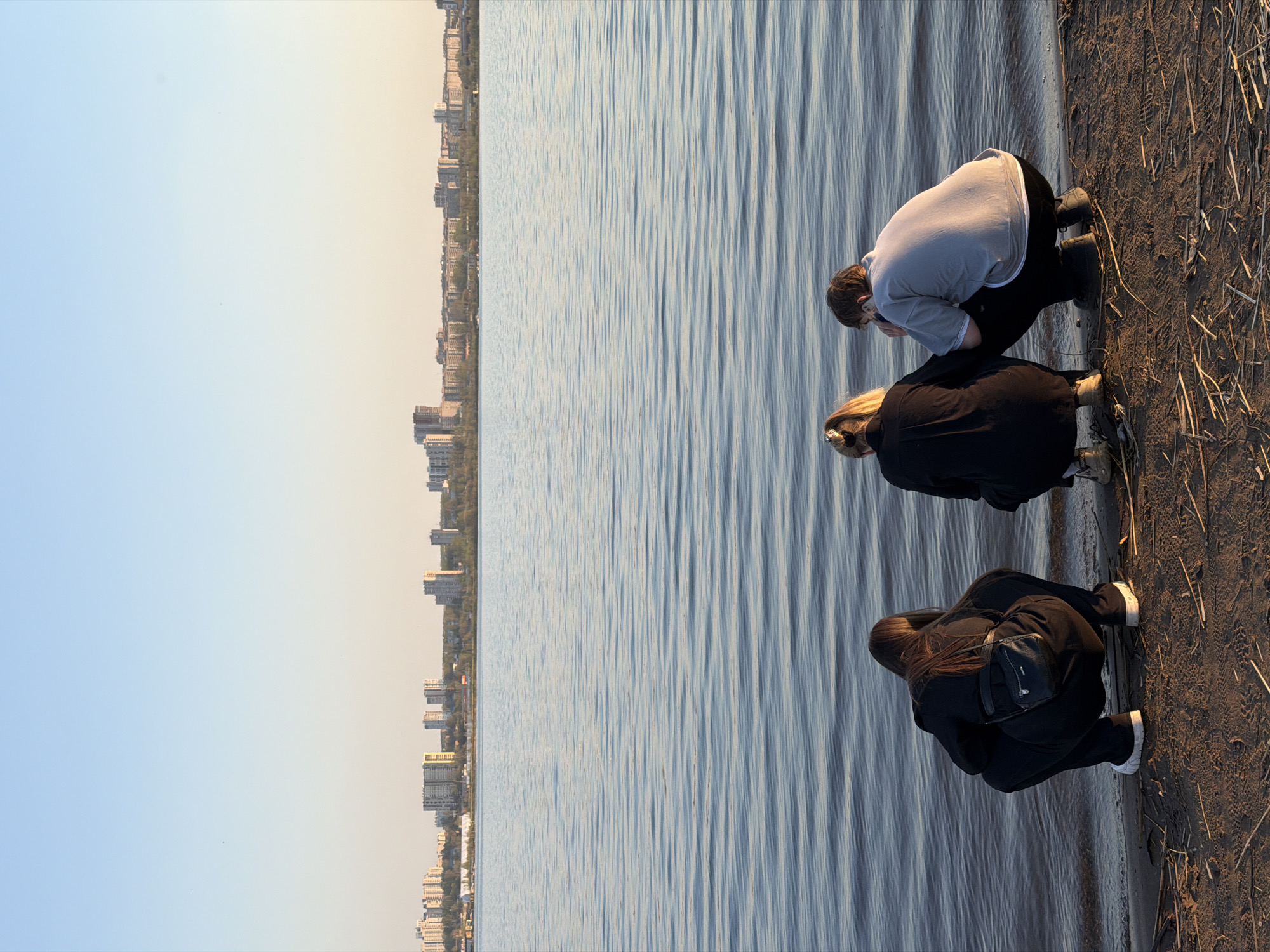 The image shows three people squatting on a shoreline next to a body of water. The person closest to the camera wears a black jacket, black pants, and white shoes, with a black bag slung over their shoulder. The person in the middle has blonde hair and wears a black jacket with beige shoes. The person farthest from the camera wears a light gray sweatshirt and black pants with black shoes and appears to be using a phone. To the left of the water, a city skyline is visible, suggesting an urban environment. The shoreline is covered with dark earth and scattered dry reeds. This location is in the city of Izhevsk, Russia.