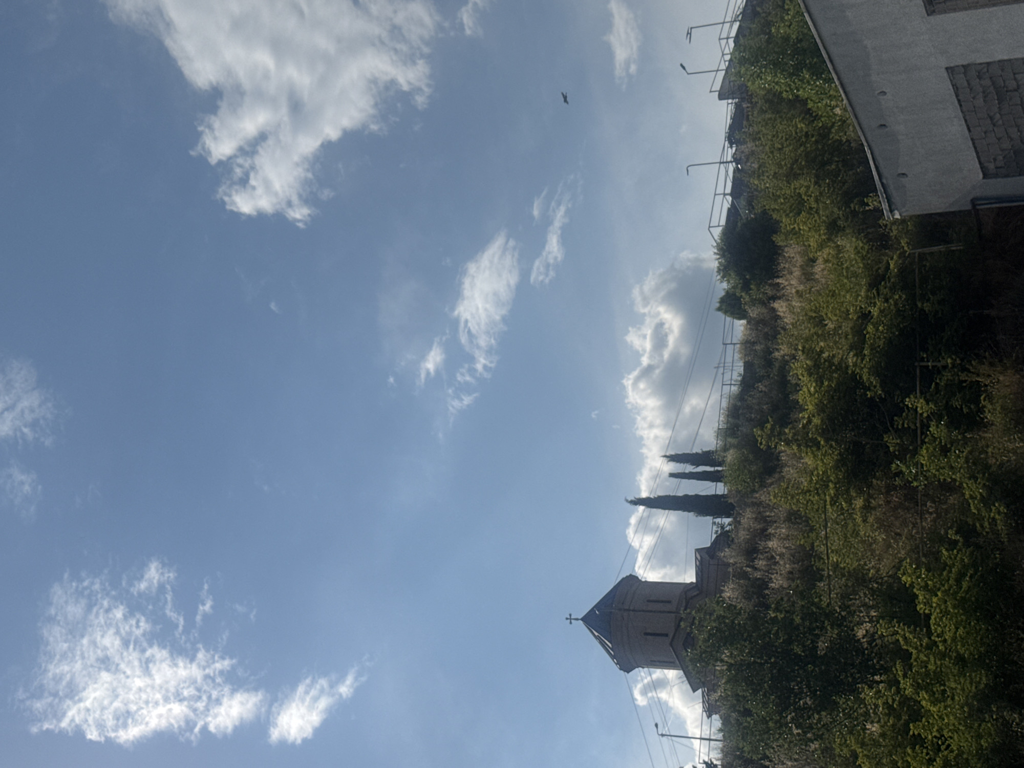 The image depicts a low-angle view of a church situated on a hill in Tbilisi, Georgia, under a bright blue sky with scattered clouds. The church has a conical roof topped with a cross, and its exterior appears to be made of light-colored stone. Several tall, slender evergreen trees stand near the church. The hill is covered in dense vegetation, including trees and shrubs. In the foreground, the roof and upper wall of a building are visible, constructed with gray siding. The overall scene suggests a sunny day in an urban environment with natural elements. A small bird is visible flying in the sky.