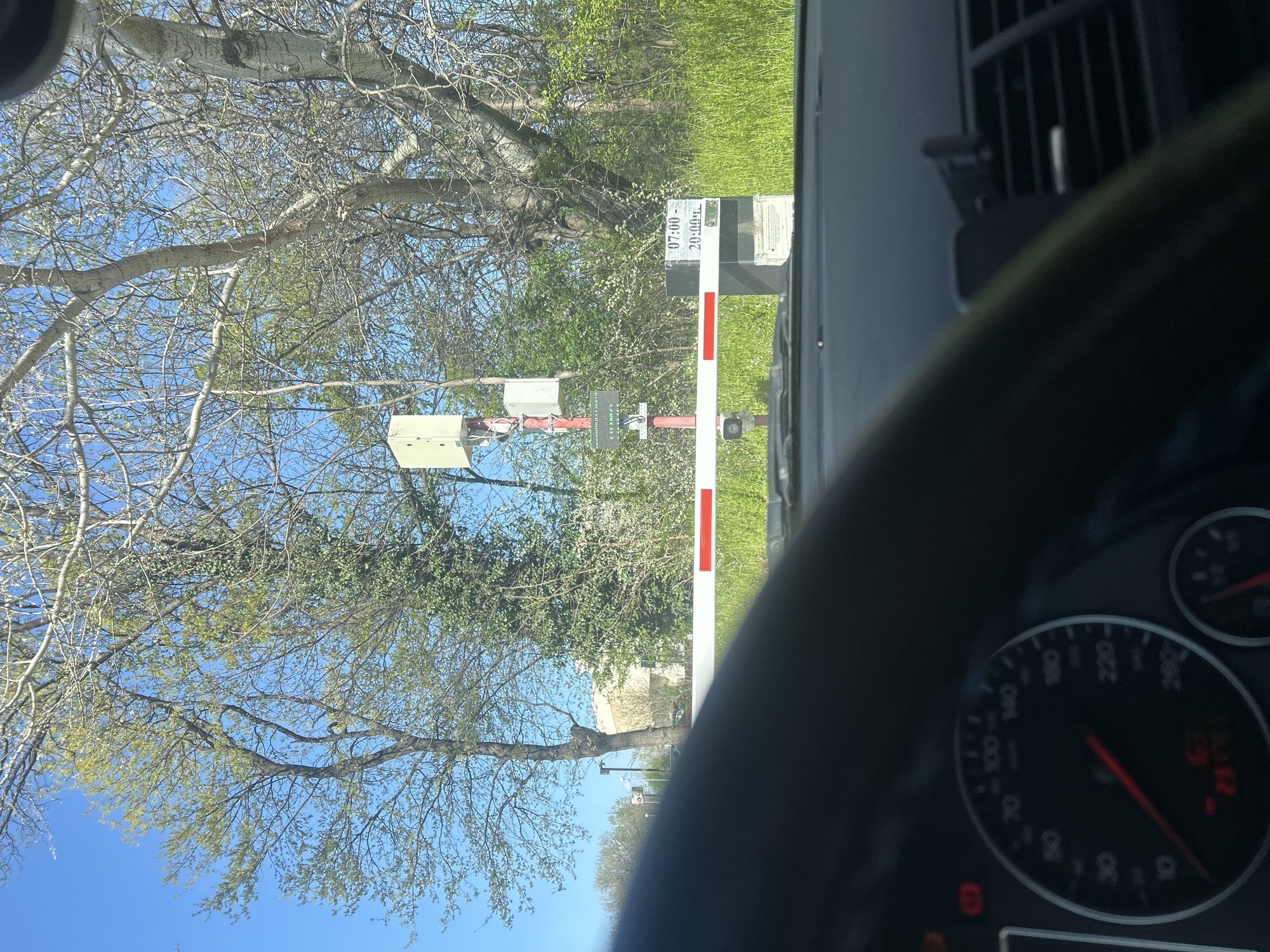 The image appears to be taken from inside a car. The viewpoint is through the steering wheel and dashboard. The car is stopped at a barrier gate. The gate is white with red horizontal stripes and a sign indicating the gate is closed between 07:00 and 20:00. Electrical or communication boxes are attached to the support structure of the gate. A small CCTV camera is also visible attached to the support. The barrier is surrounded by trees and grass. The sky is blue. Considering the location of the gate, the image is potentially taken near the Rowing Channel in Plovdiv, Bulgaria, where gated access is known.