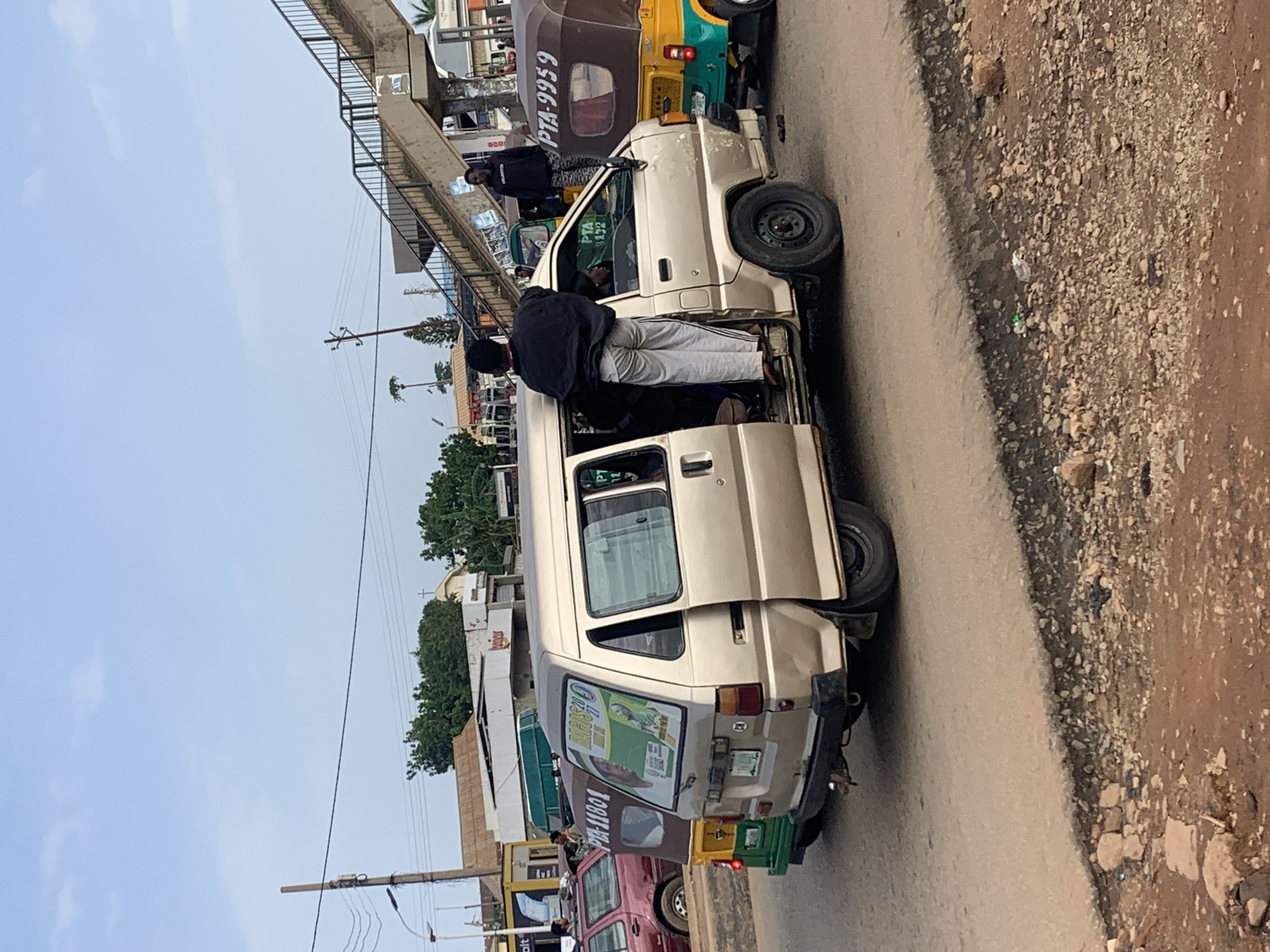 In Gura Top, Nigeria, on a clear day, a beige van is parked on a dirt road. A man in a black sweatshirt and light-colored pants stands on the open side door of the van, leaning in. Another person is visible inside the van. Behind the van, a yellow and green auto rickshaw is seen with the license plate "PTA-9959". Further in the background, a pedestrian bridge crosses over the road, and several buildings and trees are visible. Power lines and a street light are also present, indicating an urban or semi-urban setting. The road surface shows signs of wear and tear, with gravel and dirt on the side.