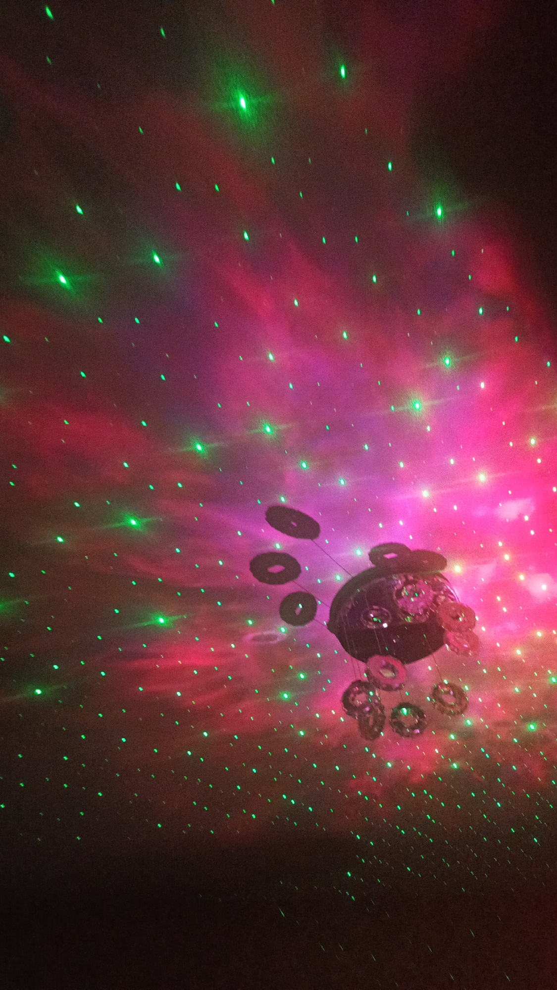 An image shows a ceiling lit by a projection of green laser dots and pink nebulae. A dark, spherical object with multiple circular, ring-shaped protrusions hangs in the center of the image. The rings cast shadows on the ceiling. The lighting suggests a room or event space. There is no discernible information about the city of Zvenigorod, Russia, within the image.
