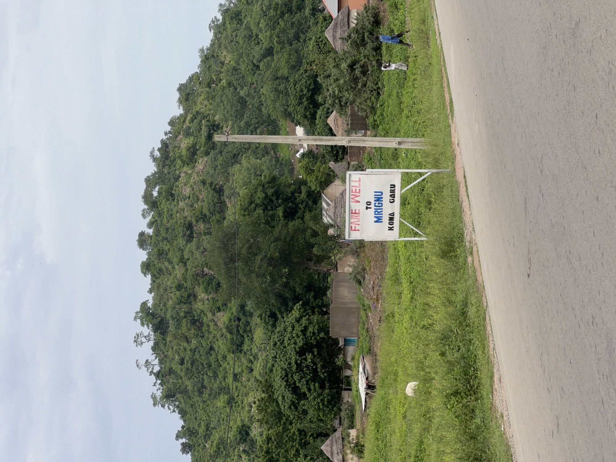 This media file depicts a rural scene in Minda, Nigeria. A paved road runs along the right side of the frame, bordered by a strip of lush green grass and vegetation. On the left, a steep, densely forested hill rises, with scattered trees and rocky outcrops visible.
In the middle ground, a white sign with blue and red lettering is prominently displayed on a metal stand. The sign reads "FATE WELL TO MRIGNU KONA GARU". Behind the sign, a utility pole stands tall, with wires extending across the scene. Several small, rustic structures with thatched roofs are nestled amongst the trees at the base of the hill, suggesting a small village or settlement.
Two figures are visible near the top of the hill, on the grassy verge beside the road. One appears to be wearing a blue garment, and the other, lighter colored clothing. Their presence suggests normal daily activity in the area. The sky is overcast, indicating a cloudy day. The overall impression is one of a quiet, natural setting with signs of human habitation. The image appears to be taken in daylight.