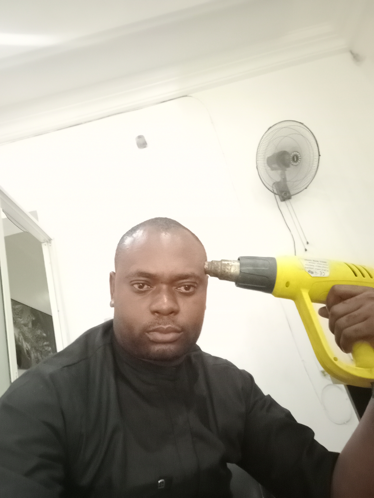 An indoor, eye-level shot in Gwarinpa, Nigeria, captures a man facing the camera while a yellow heat gun is pointed playfully at his right temple. The man has dark skin, a shaved head, and a neutral to slightly serious expression. He wears a dark, possibly black, high-collared shirt. A hand, likely holding the camera or the heat gun, is visible grasping the tool.
The background consists of plain white walls. On the right wall, a black standing fan with a circular metal grill is mounted, with wires hanging beneath it. To the left, a partial view of a doorframe or room partition is visible. The lighting is bright, suggesting an interior setting with artificial illumination. On the heat gun, some small, white text, including a "CE" mark and what appears to be "HAND WERK TOOL," is visible but mostly unreadable due to blurriness. The overall scene appears to be a staged, humorous moment.