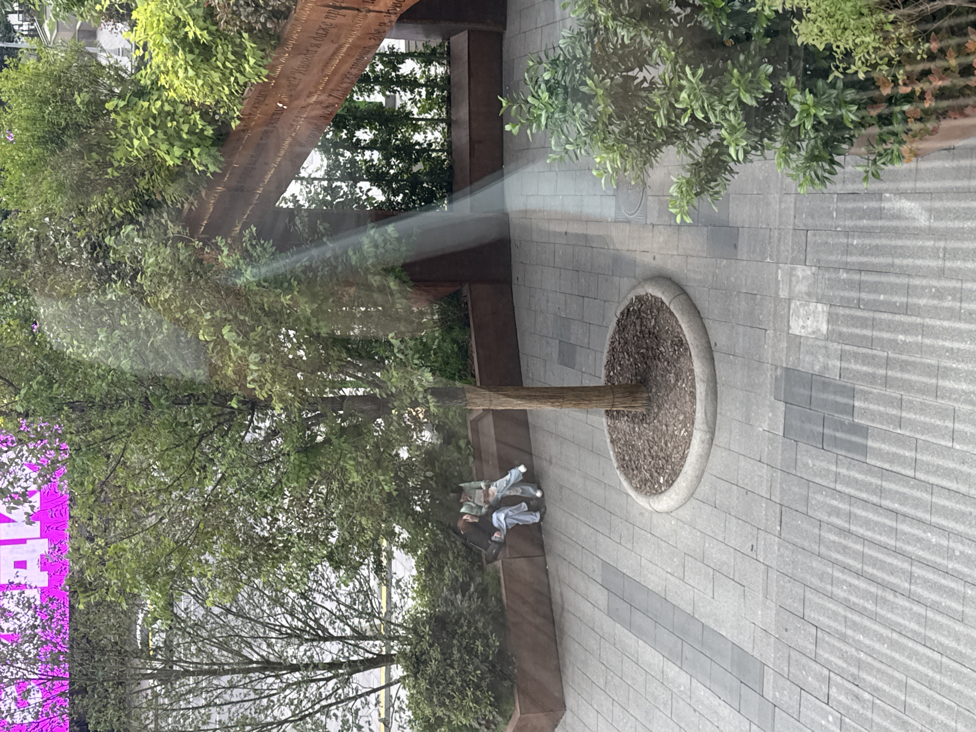 In a paved outdoor area with trees and planters, two people are sitting on a raised, dark brown bench. They appear to be reading or looking at something in their laps. One person is wearing a green top and light blue pants, while the other is wearing a dark top and light blue pants. A tree trunk rises from a circular planter filled with mulch, and its branches extend overhead, creating dappled shade. The ground is paved with grey rectangular tiles, with some darker grey tiles creating a pattern. Sunlight streams across the scene, creating bright beams. In the background, a long, dark brown metallic structure with text engraved on it is visible. A bright pink digital display with white text is also visible on the far left. The scene appears to be a public space, possibly a park or plaza, with modern landscaping. The time of day seems to be daytime, with clear or partly cloudy weather indicated by the strong sunlight and shadows. The overall mood is calm and relaxed.