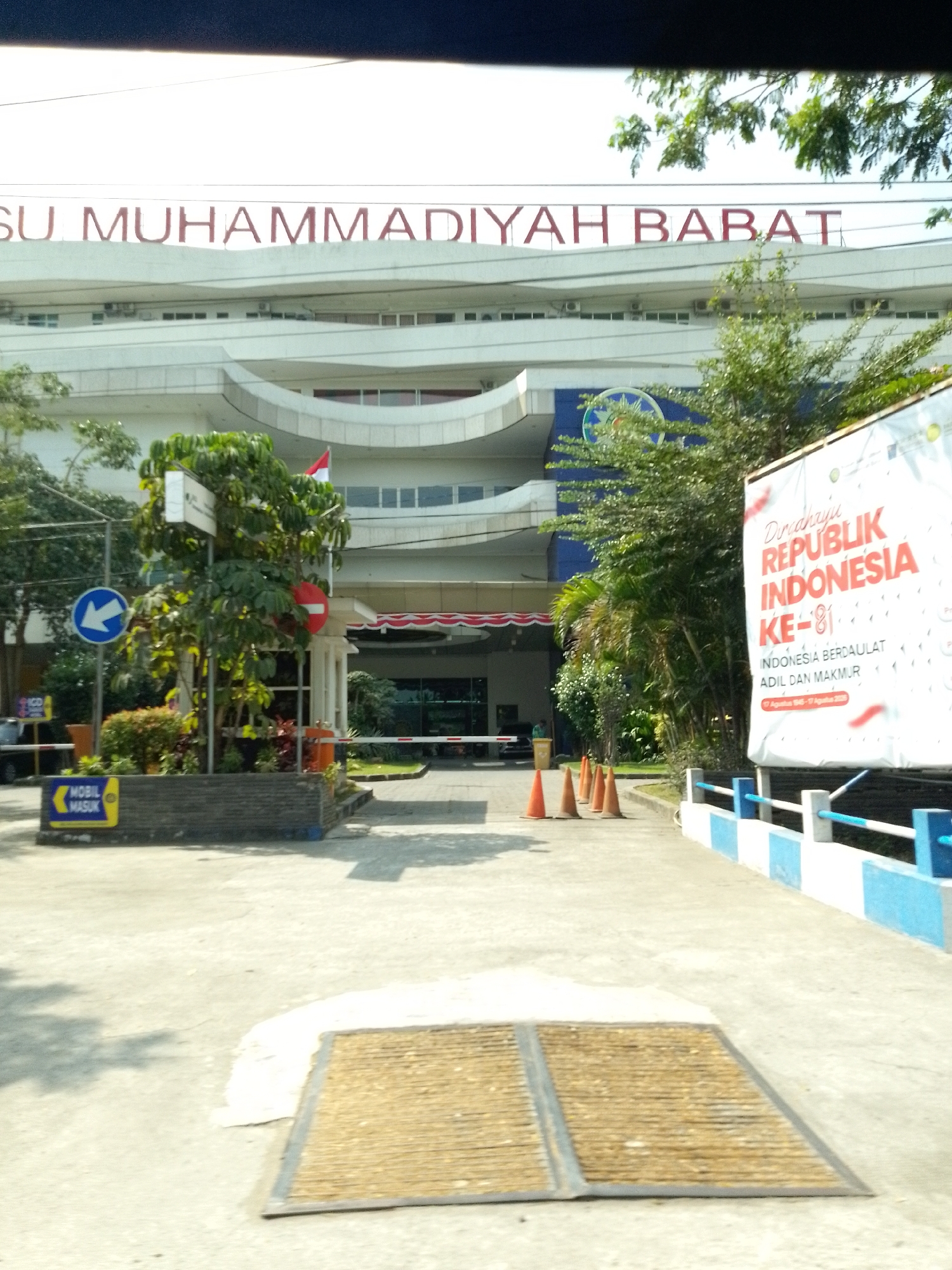 The image shows the exterior of a building with prominent red lettering spelling "SYARIF HIDAYATULLAH." The building is white with multiple stories and arched architectural elements. In front of the building, there is a concrete area with a blue and white striped railing. Several orange traffic cones are placed on the ground, suggesting traffic management or a temporary closure. To the left, there is a blue circular sign with a white arrow pointing left, indicating a direction. Below this, a yellow sign with blue text reads "MOBIL MASUK," which translates to "CAR ENTRANCE." A small structure with a barrier gate is visible. The environment appears to be an urban setting, with trees and foliage present. A large banner with text in red and white is also visible to the right, depicting Indonesian national symbols. The overall scene is a daytime outdoor shot. The location is identified as the city of Plaosan, Indonesia, specifically suggesting this is a public institution or business entrance.