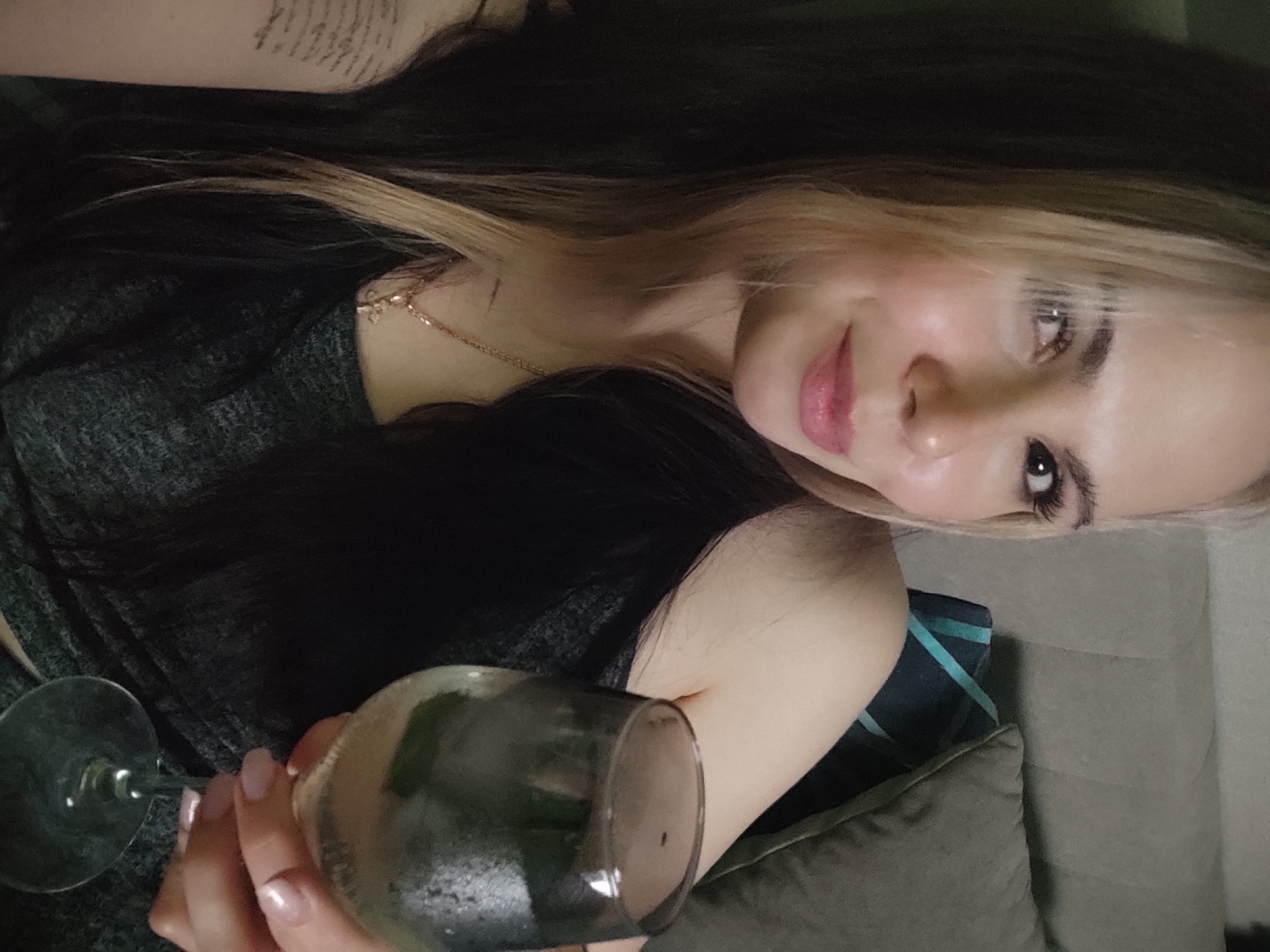A woman with two-tone blonde and black hair is holding a wine glass filled with a beverage and ice. She is wearing a gray top and a gold necklace. The background shows a pillow and a partial view of a wall. The exact location in the city of Chaikovsky, Russia, cannot be determined from the image.