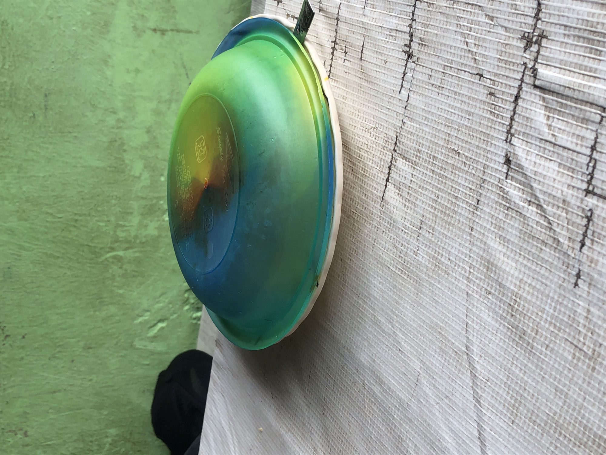 A close-up, eye-level shot shows a colorful plastic bowl resting on a textured, light-colored surface. The bowl, predominantly green and blue with a gradient effect, is upside down, revealing text and markings on its underside. It sits on a white plate. Behind the bowl, a green, textured wall is visible, with a black object, possibly a hat, placed on the left side of the frame. The scene appears to be outdoors, in Kabong, Nigeria, potentially a market or street setting, given the rough texture of the surface and the simple backdrop. The lighting suggests it is daytime, with sunlight casting shadows.