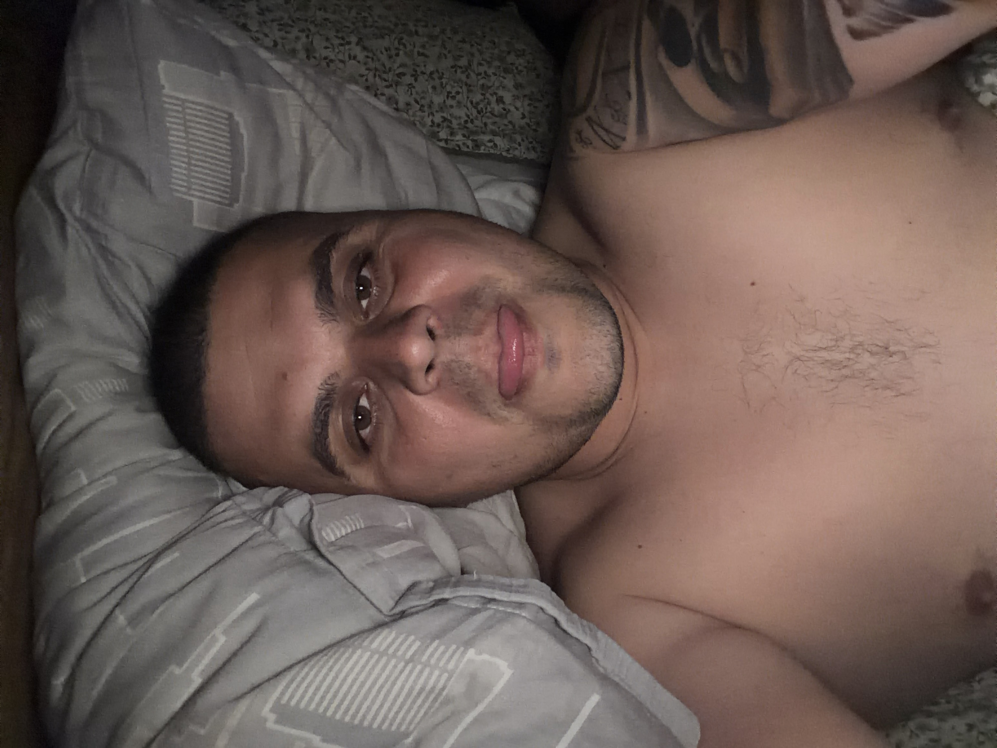 A man is depicted lying on his side in what appears to be a dimly lit bedroom. He is shirtless, facing the camera with his head resting on a light gray pillow that features a subtle geometric pattern of lines and rectangles. The man has short, dark hair, brown eyes, and visible stubble or a short beard. He is looking directly at the camera with his tongue slightly extended, suggesting a relaxed or playful expression. A prominent dark tattoo covers part of his left arm and shoulder. The bedding behind him includes a darker, possibly patterned sheet or blanket. The overall lighting is subdued, indicating either nighttime or a room with low illumination. No text is visible in the image. The provided location context for this scene is Shumen, Bulgaria.