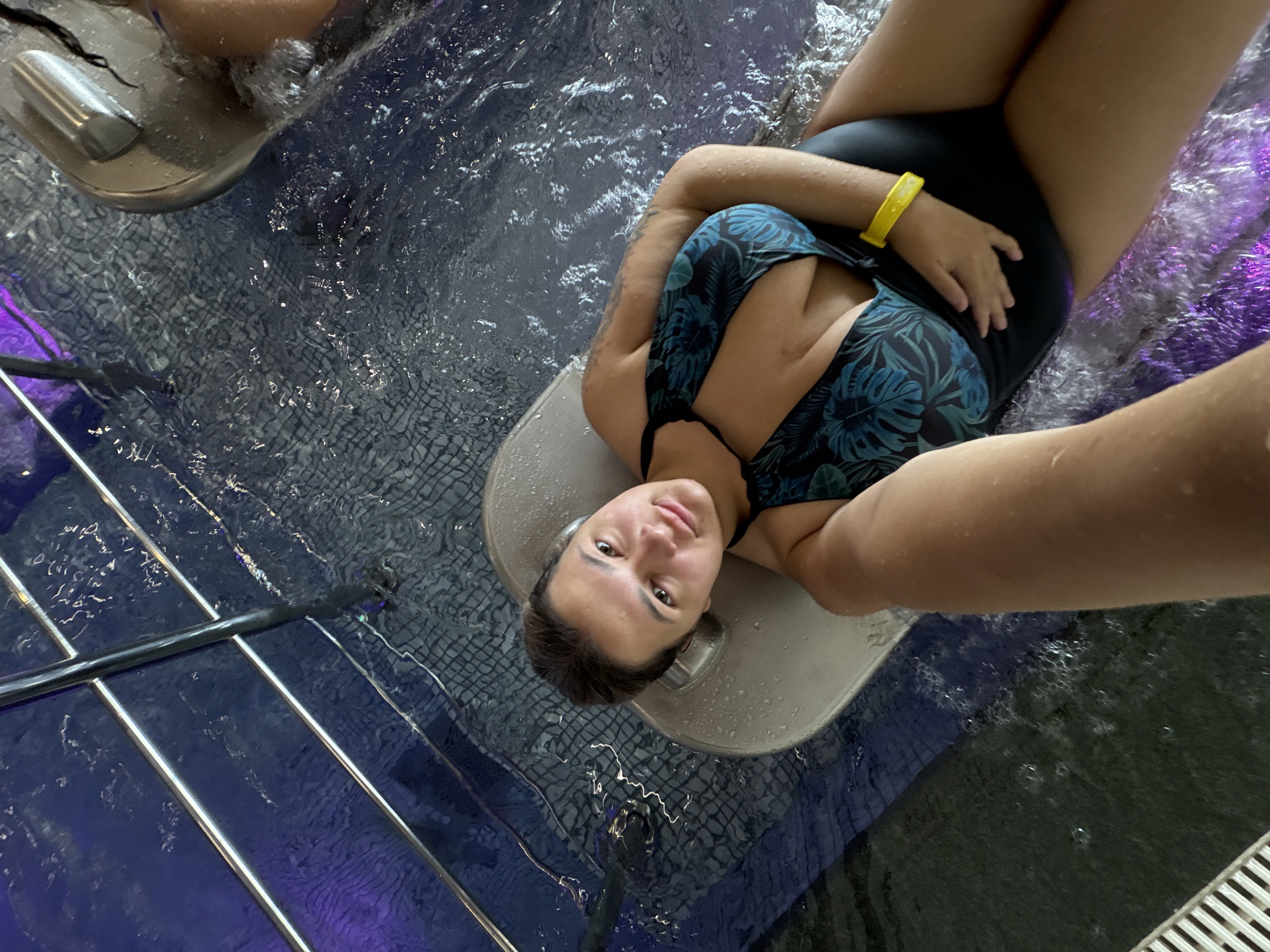 A young woman in a black and teal floral swimsuit reclines in a spa. She has her head resting on a light-colored inflatable headrest, and her right arm is bent, with her hand resting on her abdomen. A yellow bracelet is visible on her left wrist. The water around her is rippling, and there is a dark, textured surface beneath the water, possibly a tiled floor. A metal railing and blue tiled walls are visible in the background, with purple lighting adding to the ambiance. The image appears to be a selfie taken from a high angle.
